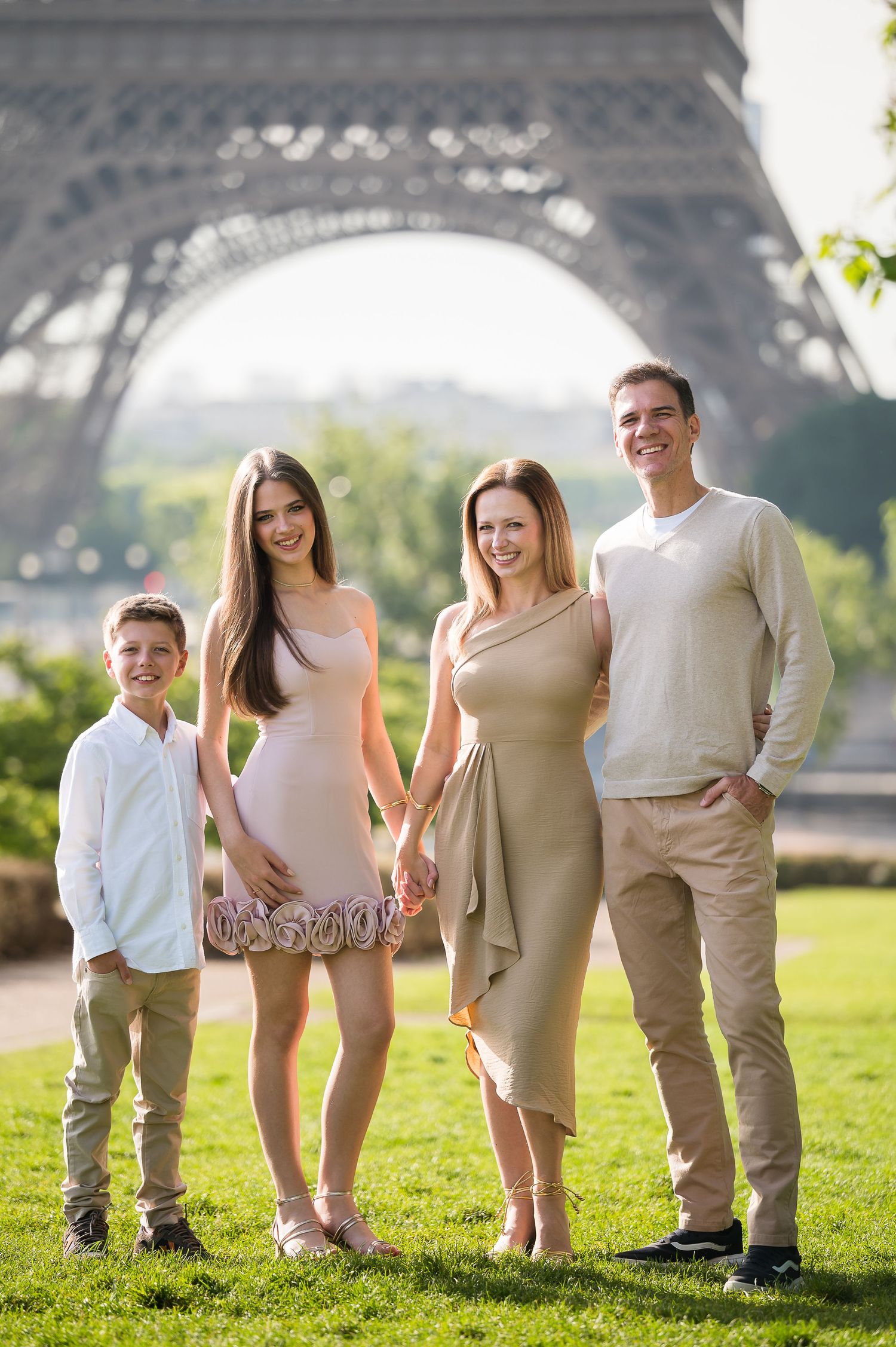 Happy family of four standing on grass with the Eiffel Tower base in the background in Paris.