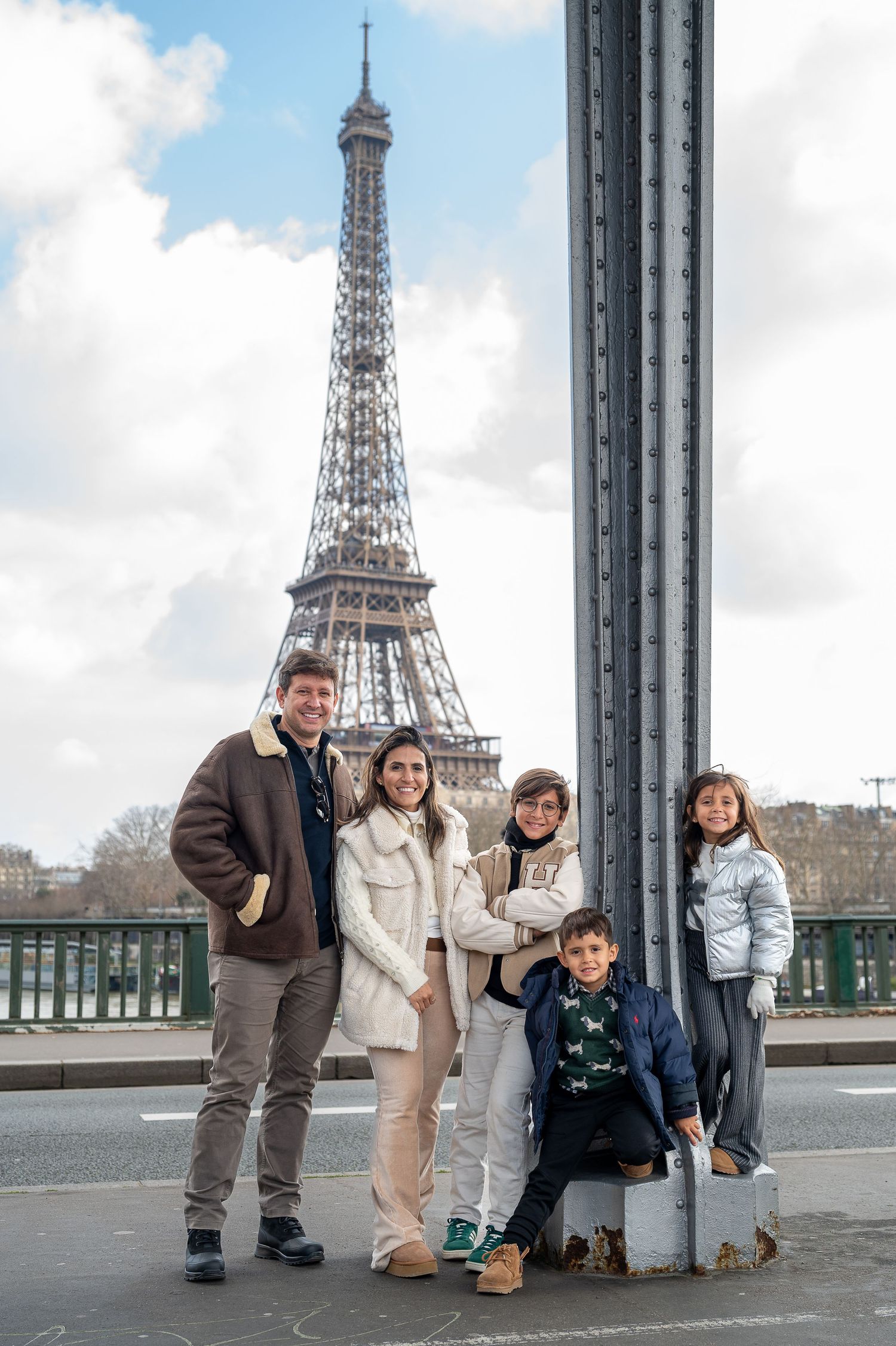 A family group poses by metal railings with the Eiffel Tower visible in the cloudy Parisian sky.