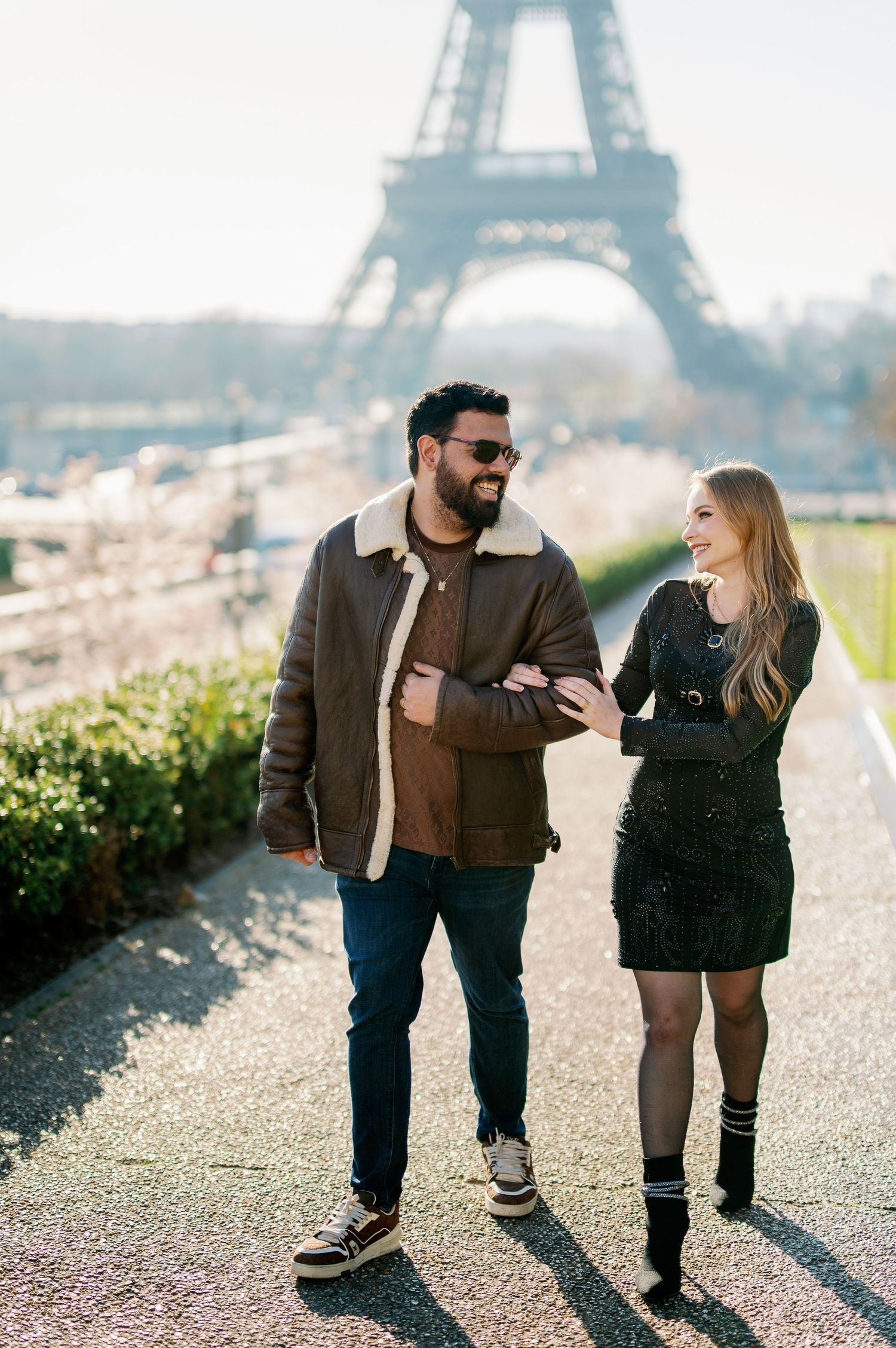 A stylish couple walks and talks together near the iconic Eiffel Tower in Paris on a sunny day.