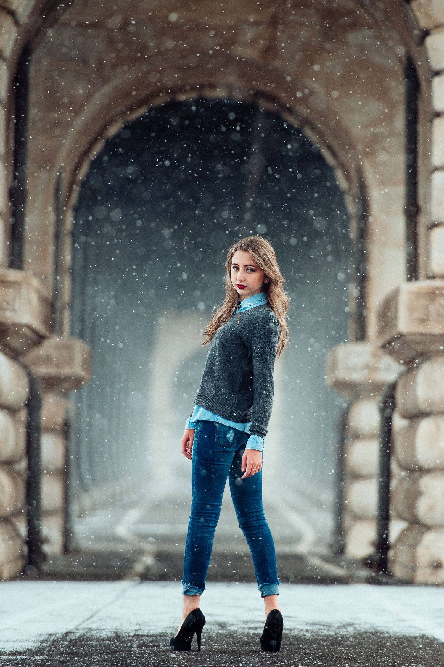 A stylish winter fashion photo taken in a stone archway while snow falls in the background.
