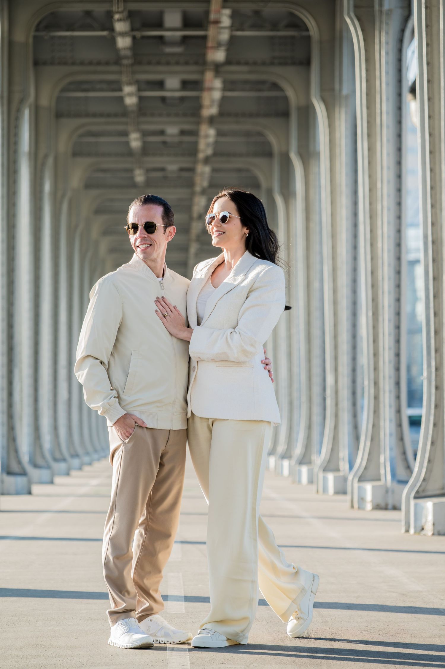 Stylish couple in white outfits and sunglasses pose together under architectural columns on a sunny day.