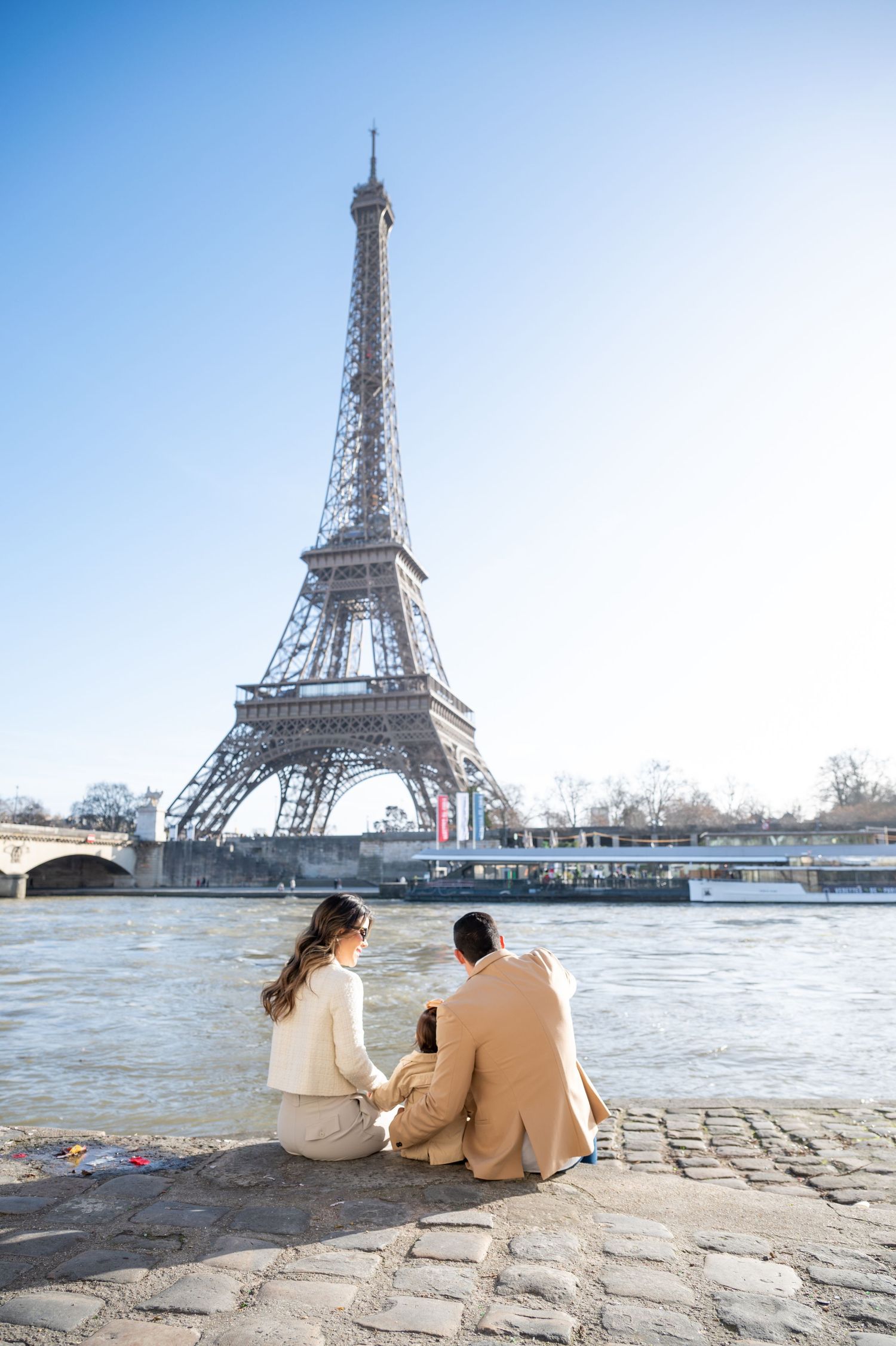 A couple sits along the Seine River overlooking the iconic Eiffel Tower on a clear day in Paris.