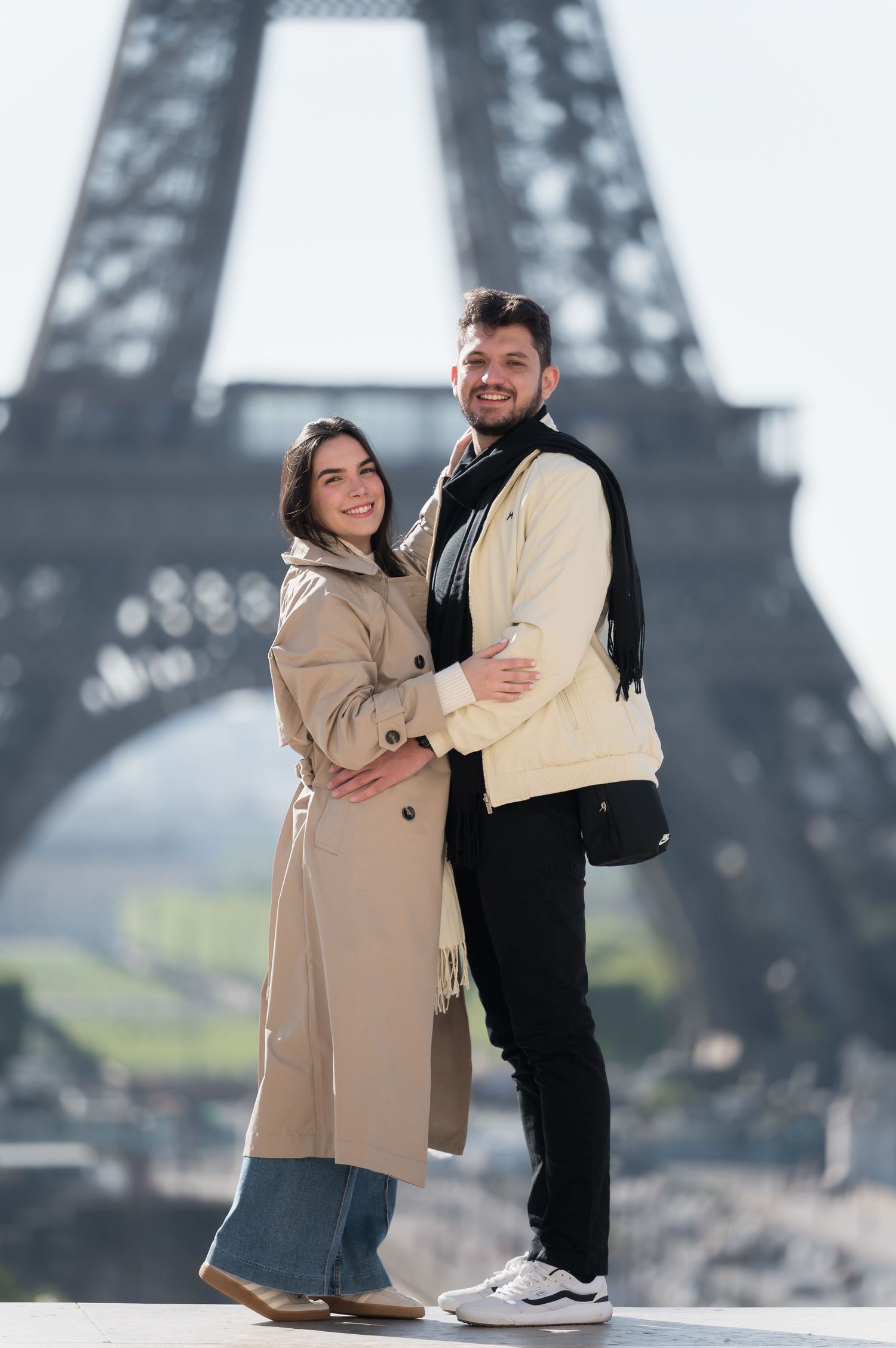 Smiling couple embracing in front of the Eiffel Tower in Paris, wearing stylish trench coats on a sunny day.