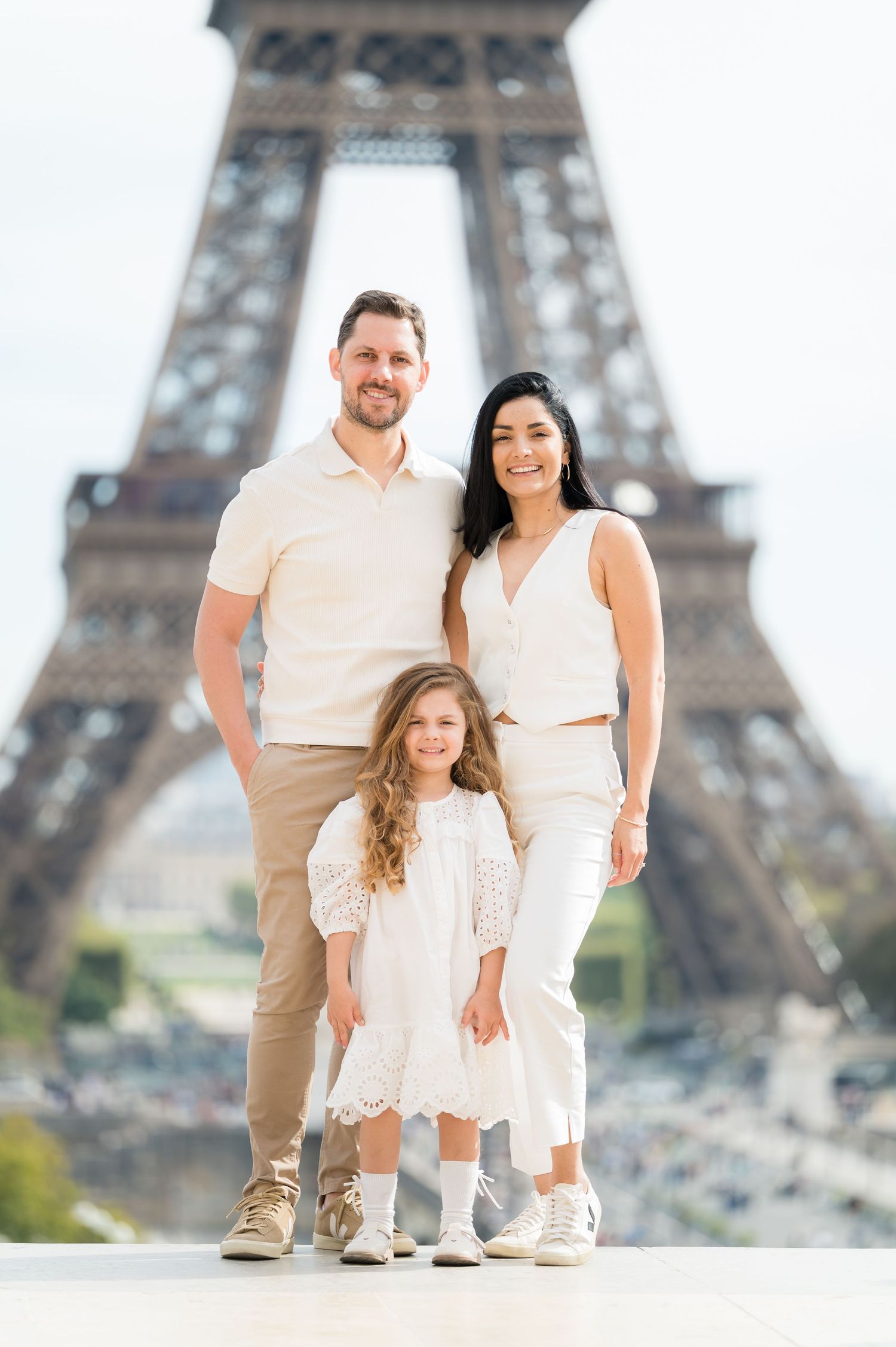 A family poses together in coordinated white and beige outfits with the iconic Eiffel Tower in Paris as their backdrop.