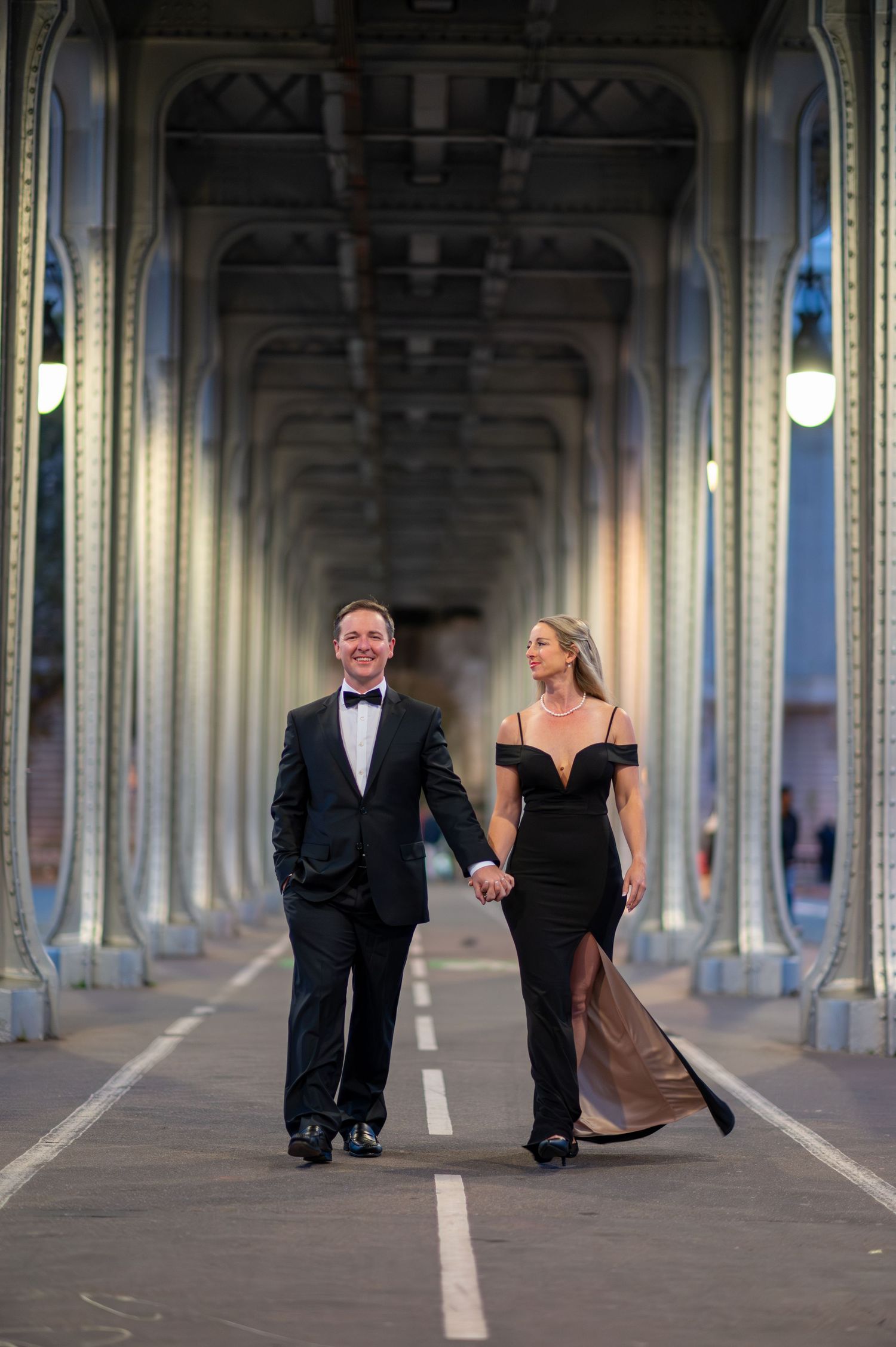 A couple in formal evening wear walks hand in hand beneath the arched colonnade of Pont de Bir-Hakeim bridge in Paris.