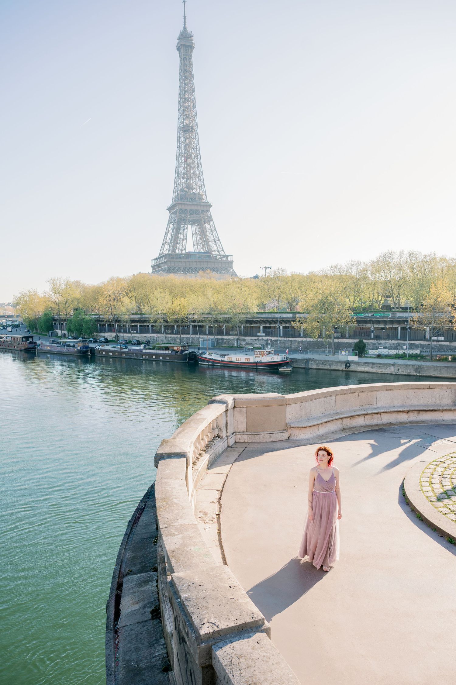 A person in a mauve dress stands along the Seine River in Paris with the iconic Eiffel Tower visible in the background.