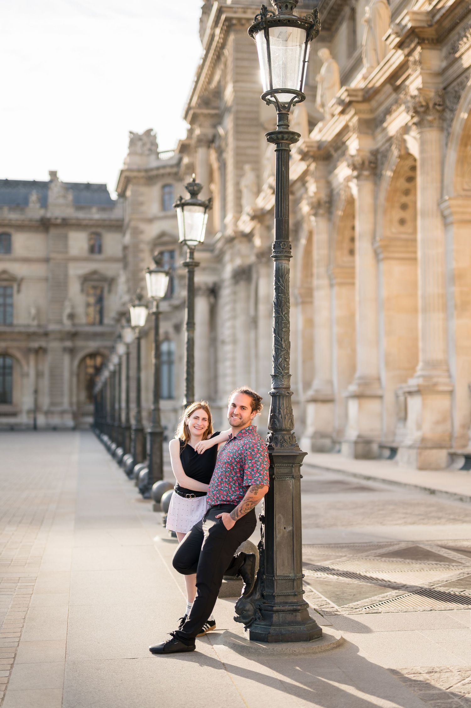 Couple posing by ornate lamp posts outside the Louvre Museum in Paris on a sunny day.