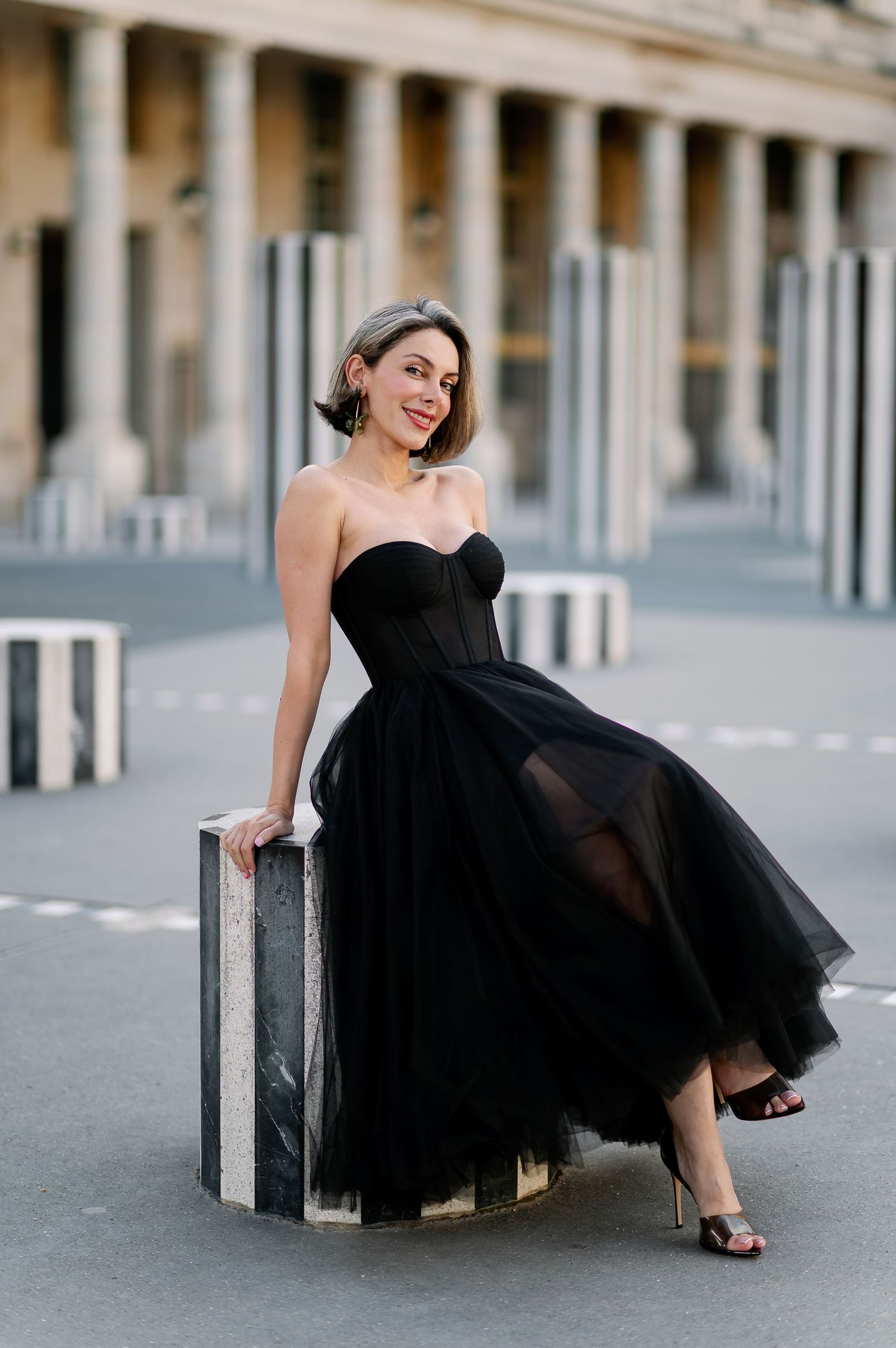 Woman in elegant black strapless tulle gown posing confidently outdoors near classic columns.