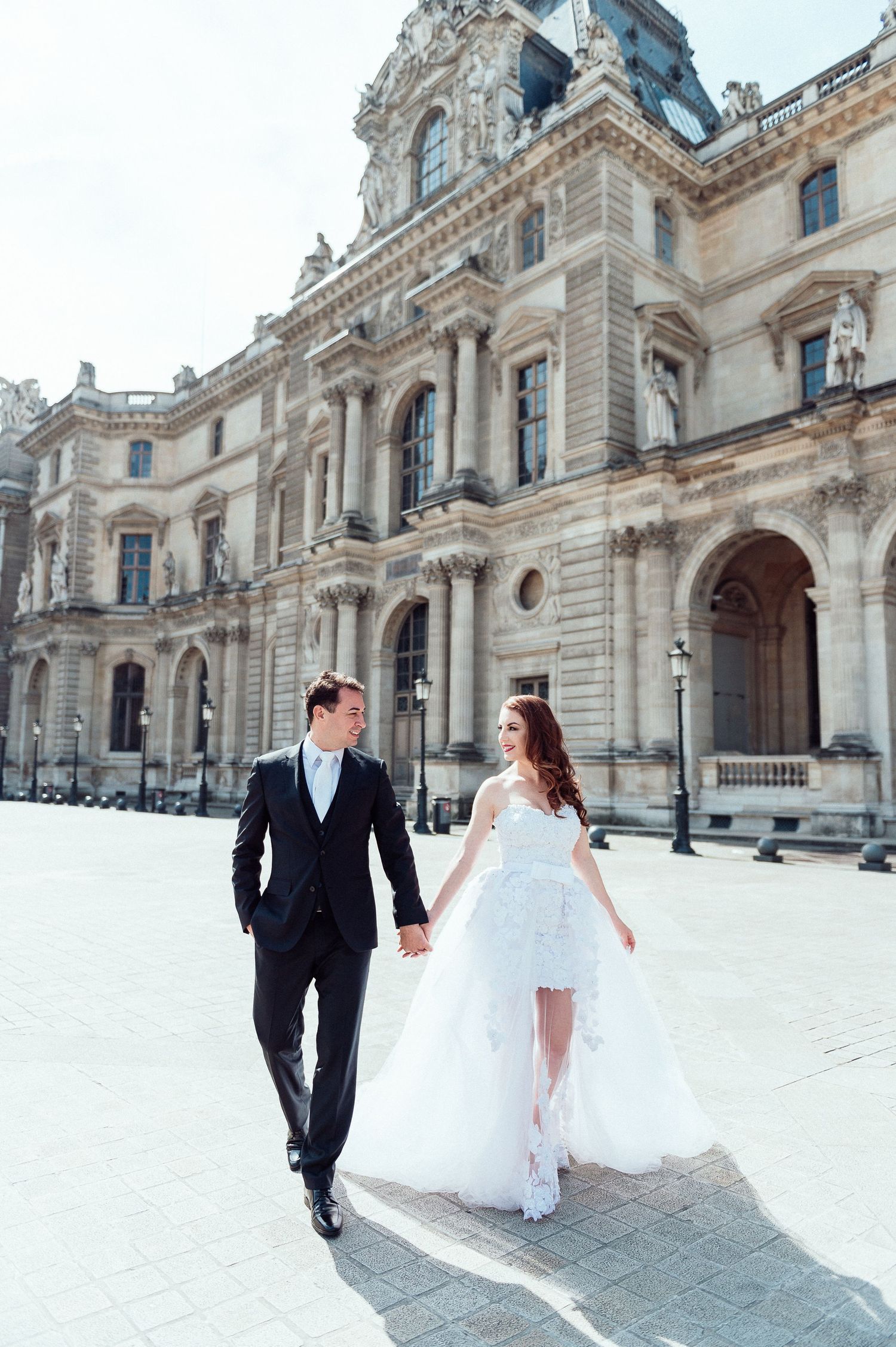 A couple walks hand in hand outside an ornate French palace building in their wedding attire.
