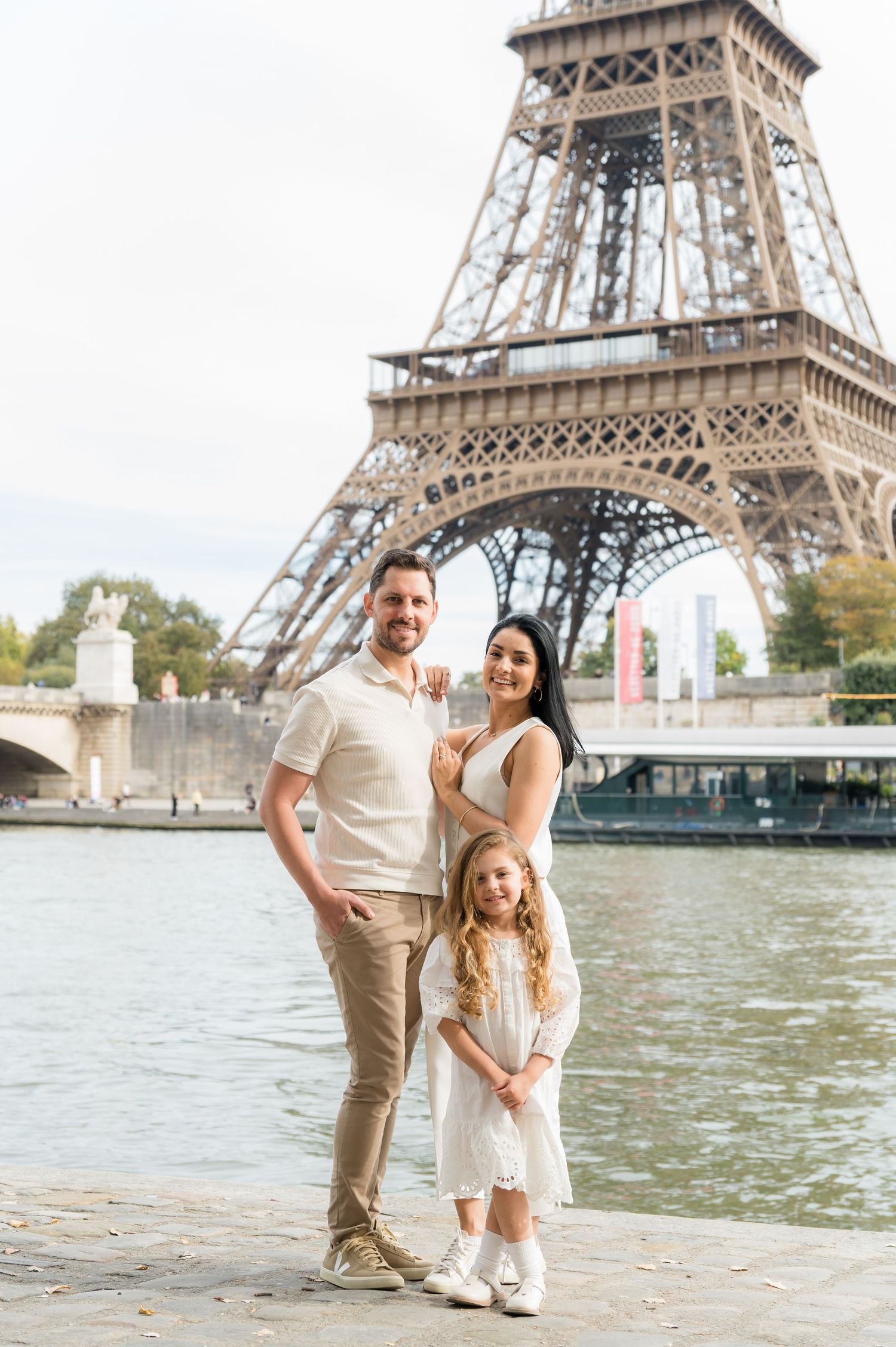 A family poses together in coordinated white outfits near the Eiffel Tower along the Seine River in Paris.