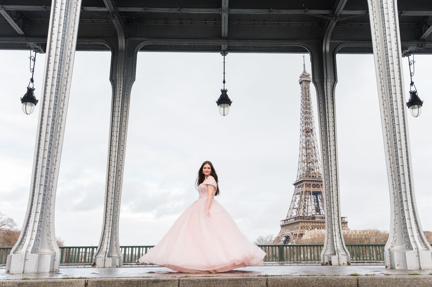 A bride in a pink ballgown poses elegantly between white columns with the Eiffel Tower visible in the background.