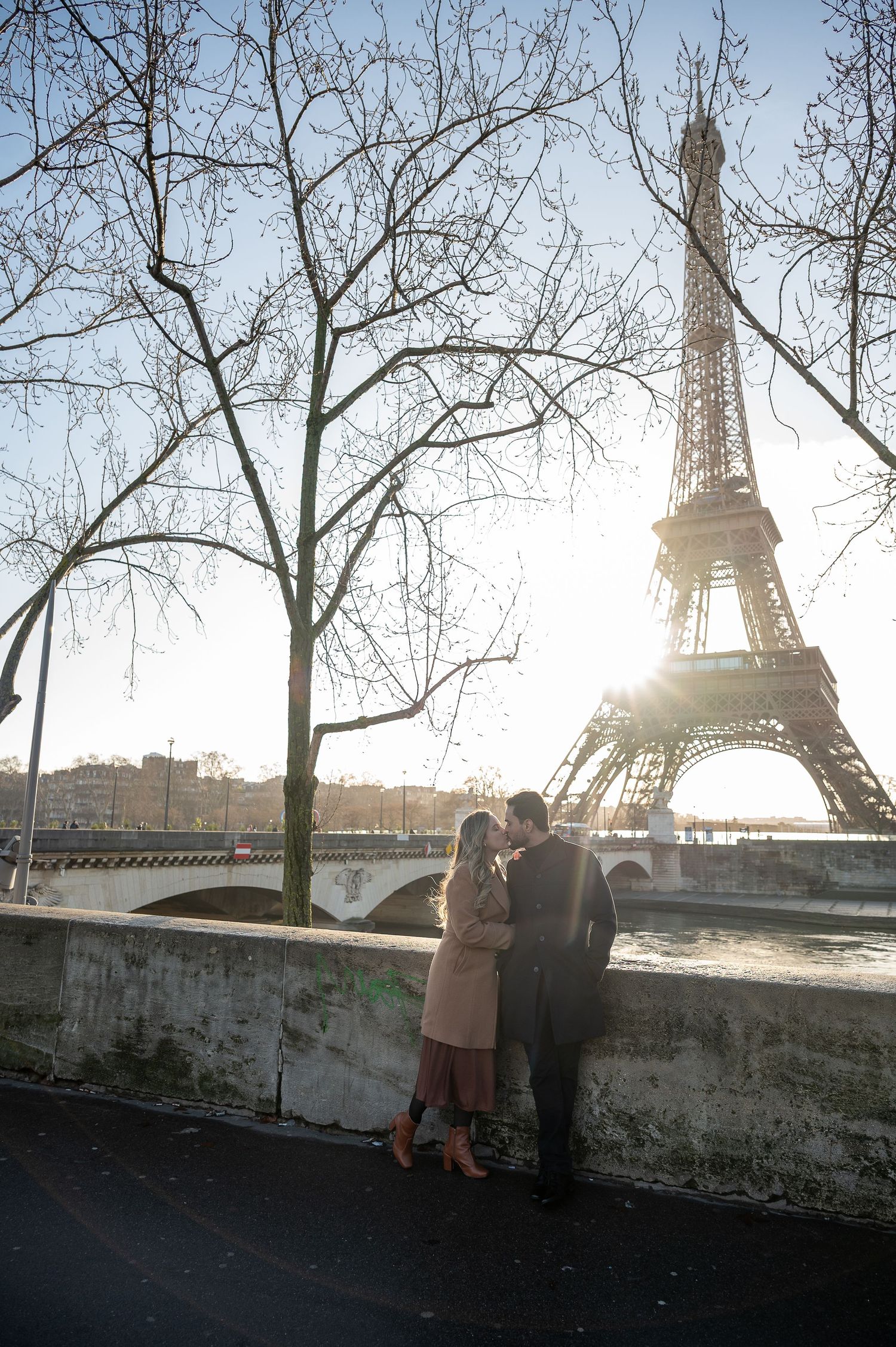 A winter scene in Paris with bare trees framing a couple and the Eiffel Tower at sunset.