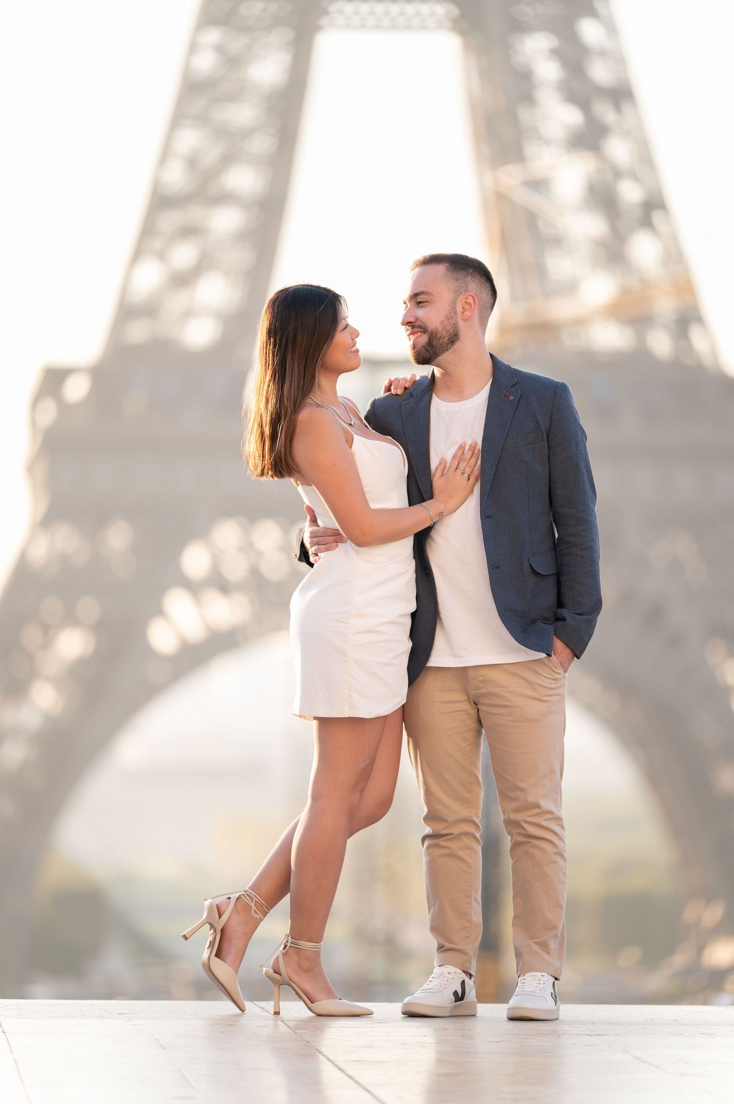 Couple posing romantically in front of the Eiffel Tower in Paris, woman in white dress, man in blue blazer.