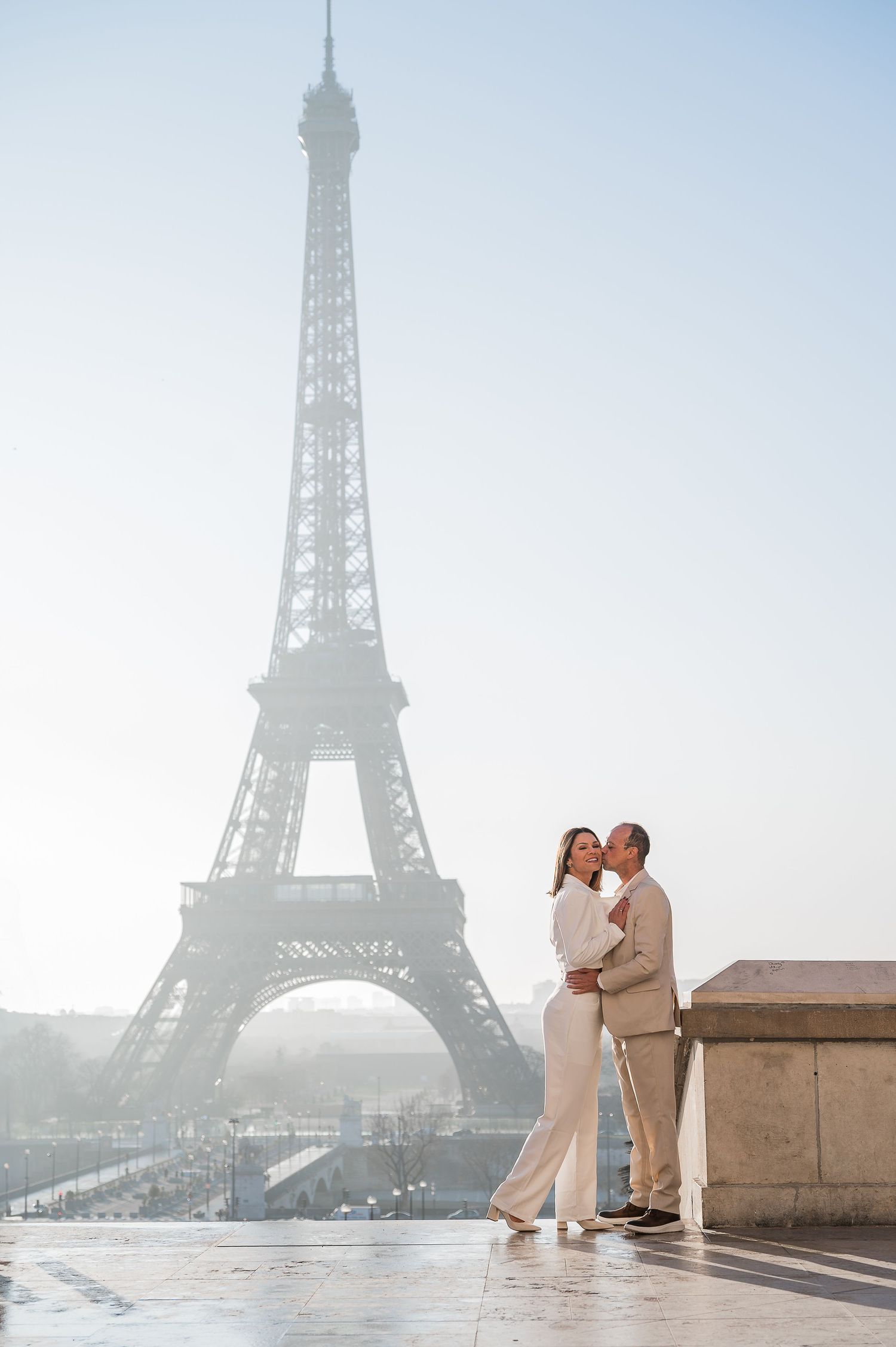 A couple embracing on a Paris viewing platform with the Eiffel Tower looming in the foggy background.