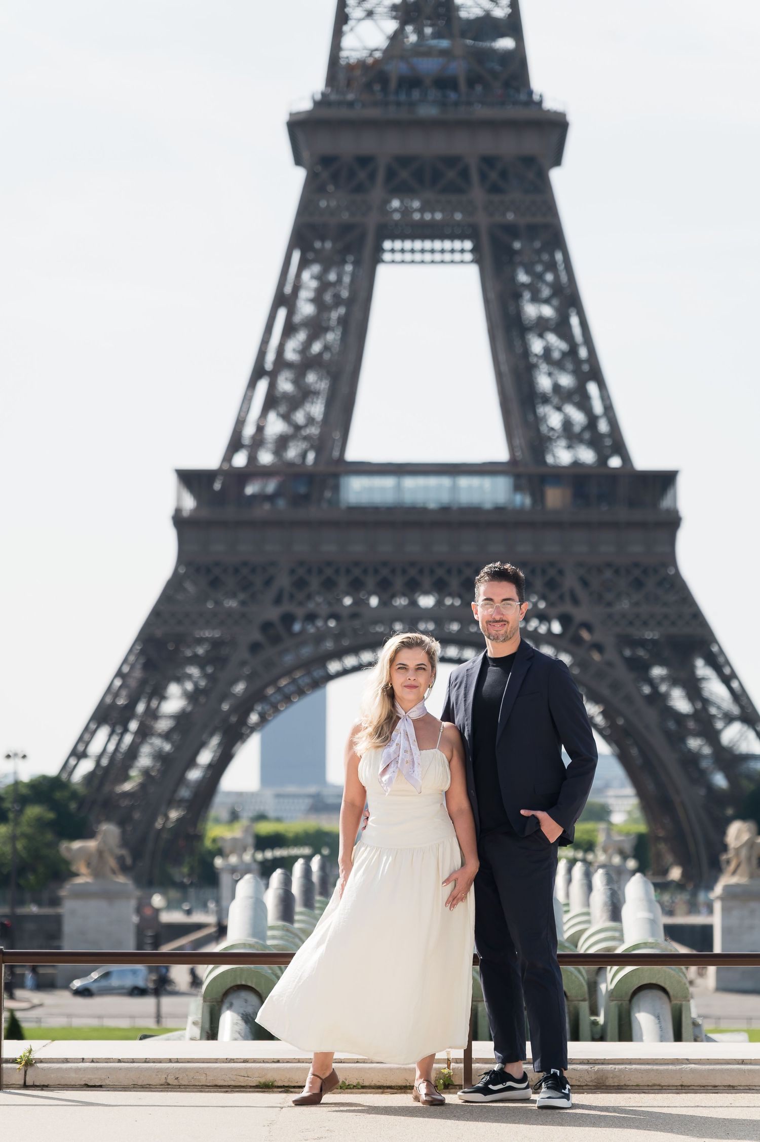 Elegant couple posing in front of the Eiffel Tower in Paris, woman in white dress, man in black outfit.