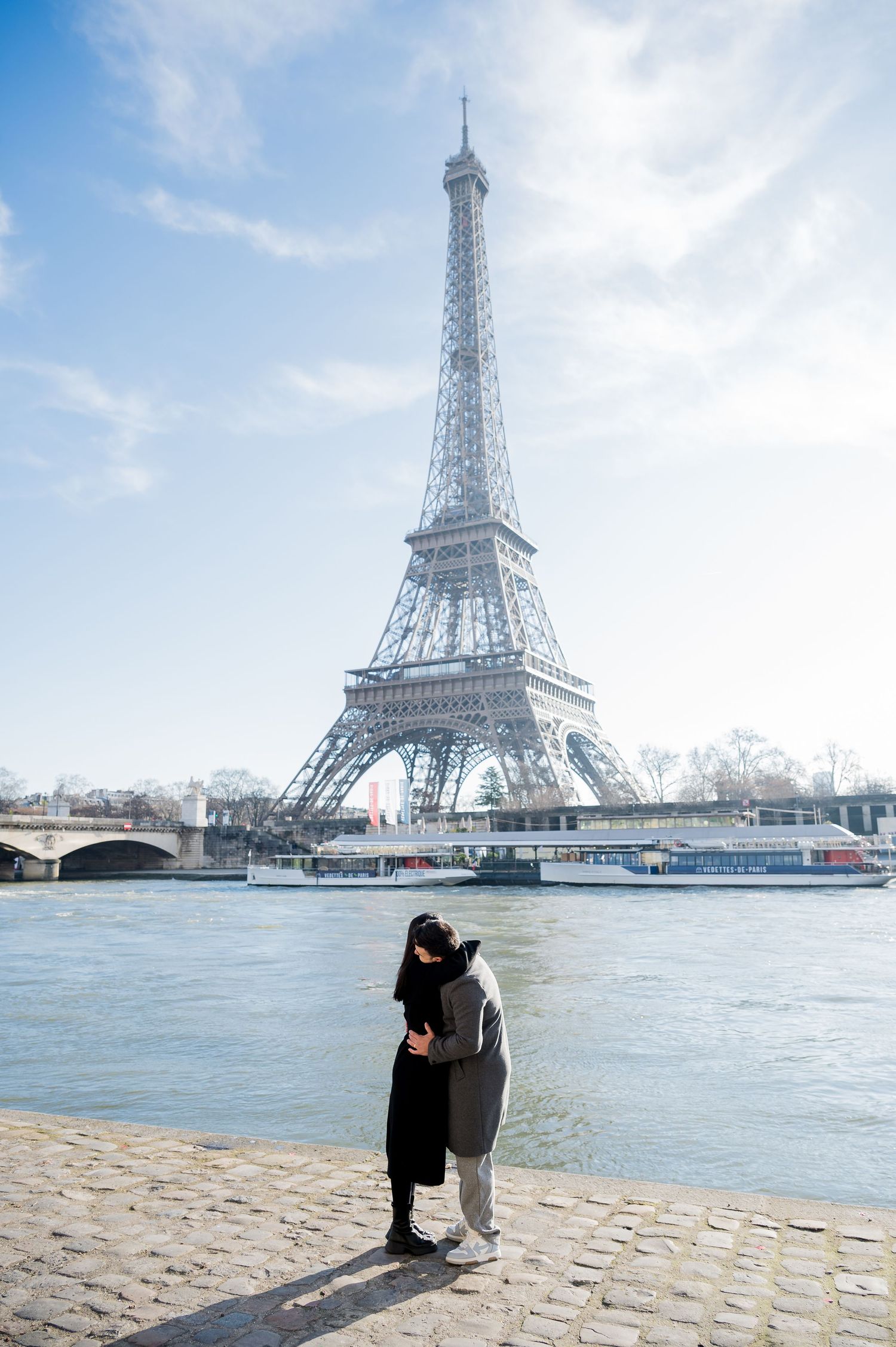 A couple embraces by the Seine River with the Eiffel Tower rising majestically in the background.