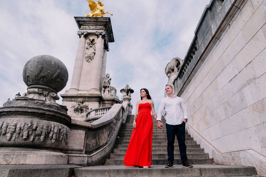 Couple standing on ornate stone steps near the Alexandre III bridge in Paris with architectural details and cloudy sky.