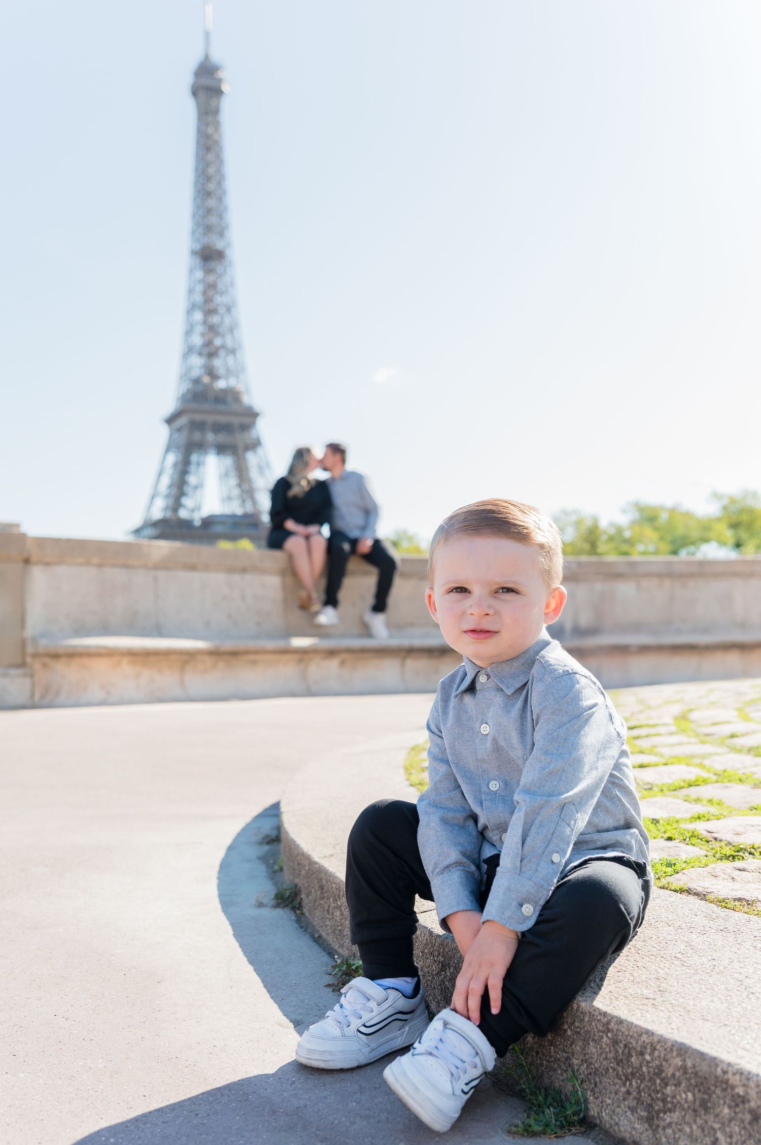 Young child in denim shirt sits casually on stone ledge with iconic Eiffel Tower visible in background.