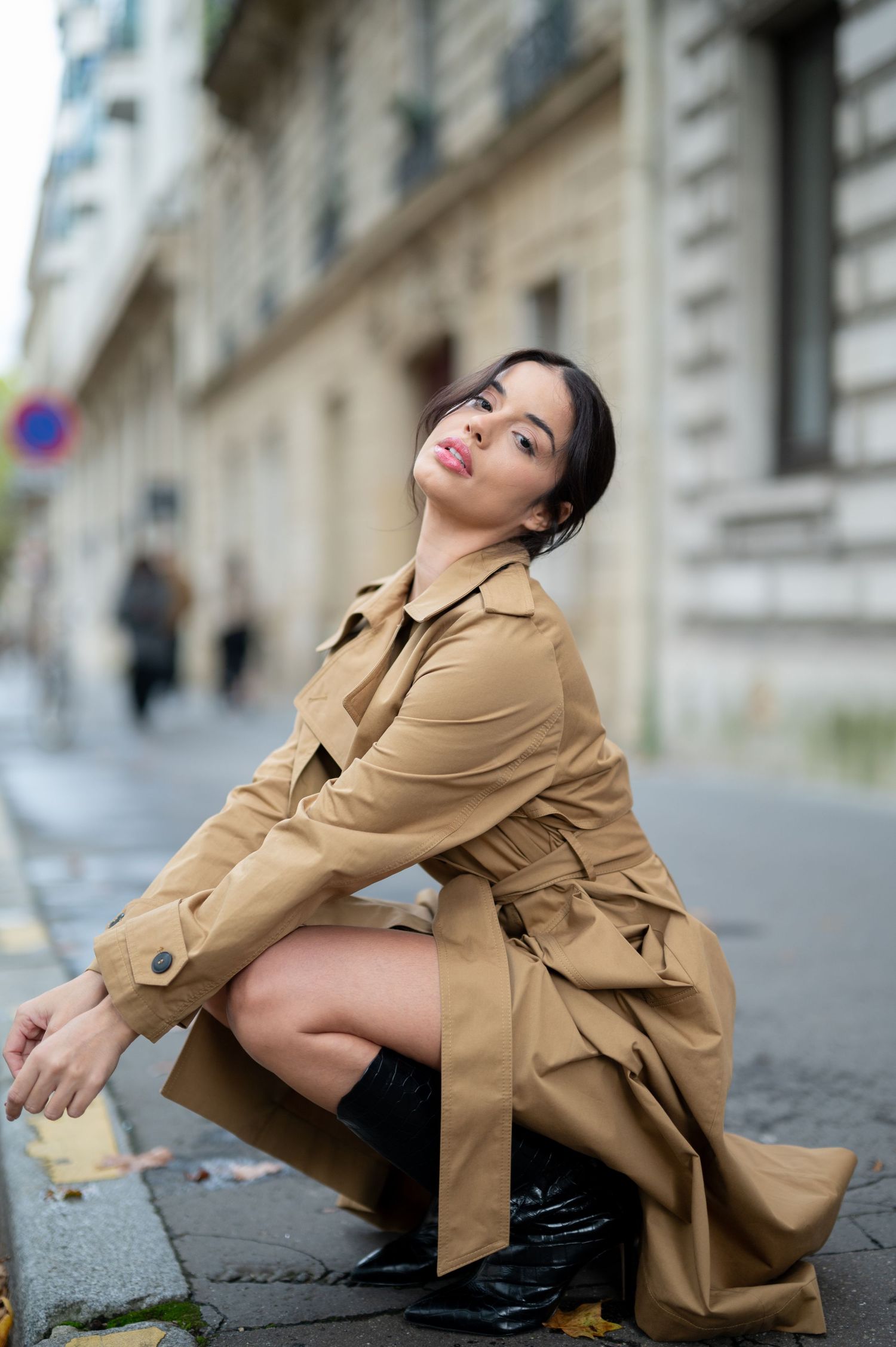 Someone wearing a tan trench coat and black boots poses on a cobblestone street in front of historic buildings.