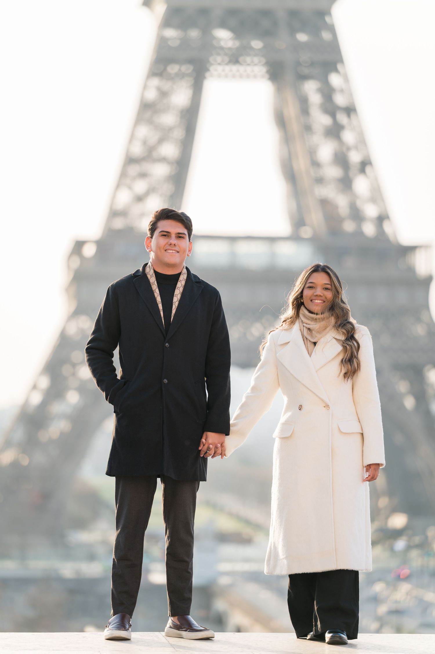 Couple holding hands in front of the Eiffel Tower in Paris, wearing elegant black and white coats.