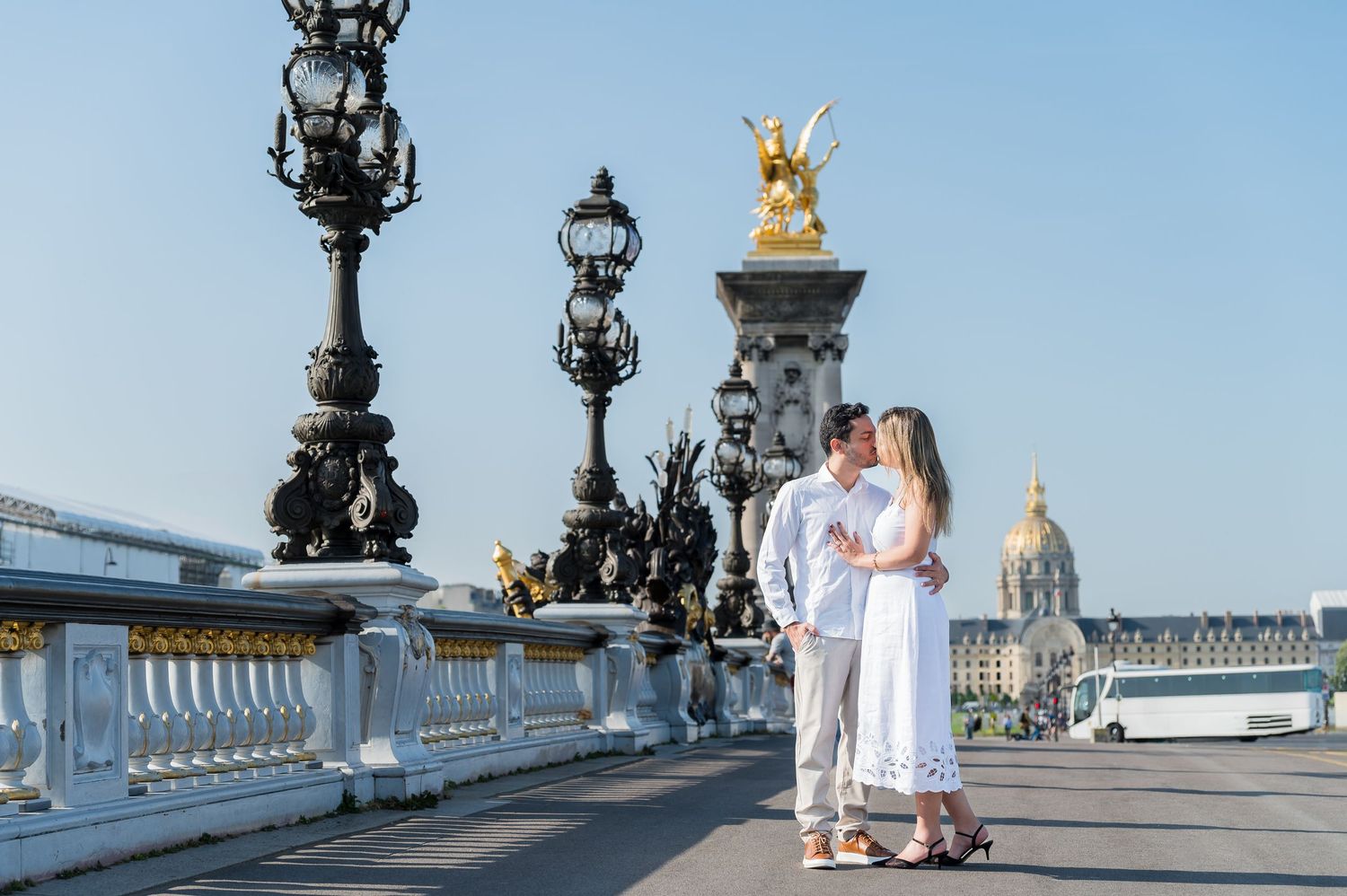 In white attire, people stroll along scenic Pont Alexandre III bridge in Paris with ornate decorative details and sky background.
