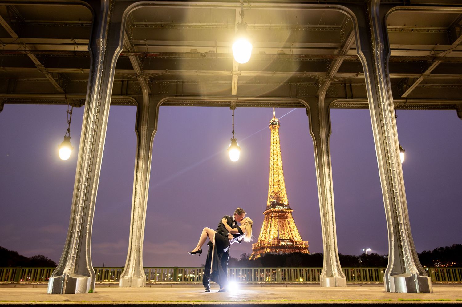 A silhouetted couple embraces beneath the Bir-Hakeim bridge in Paris with the illuminated Eiffel Tower glowing at dusk.