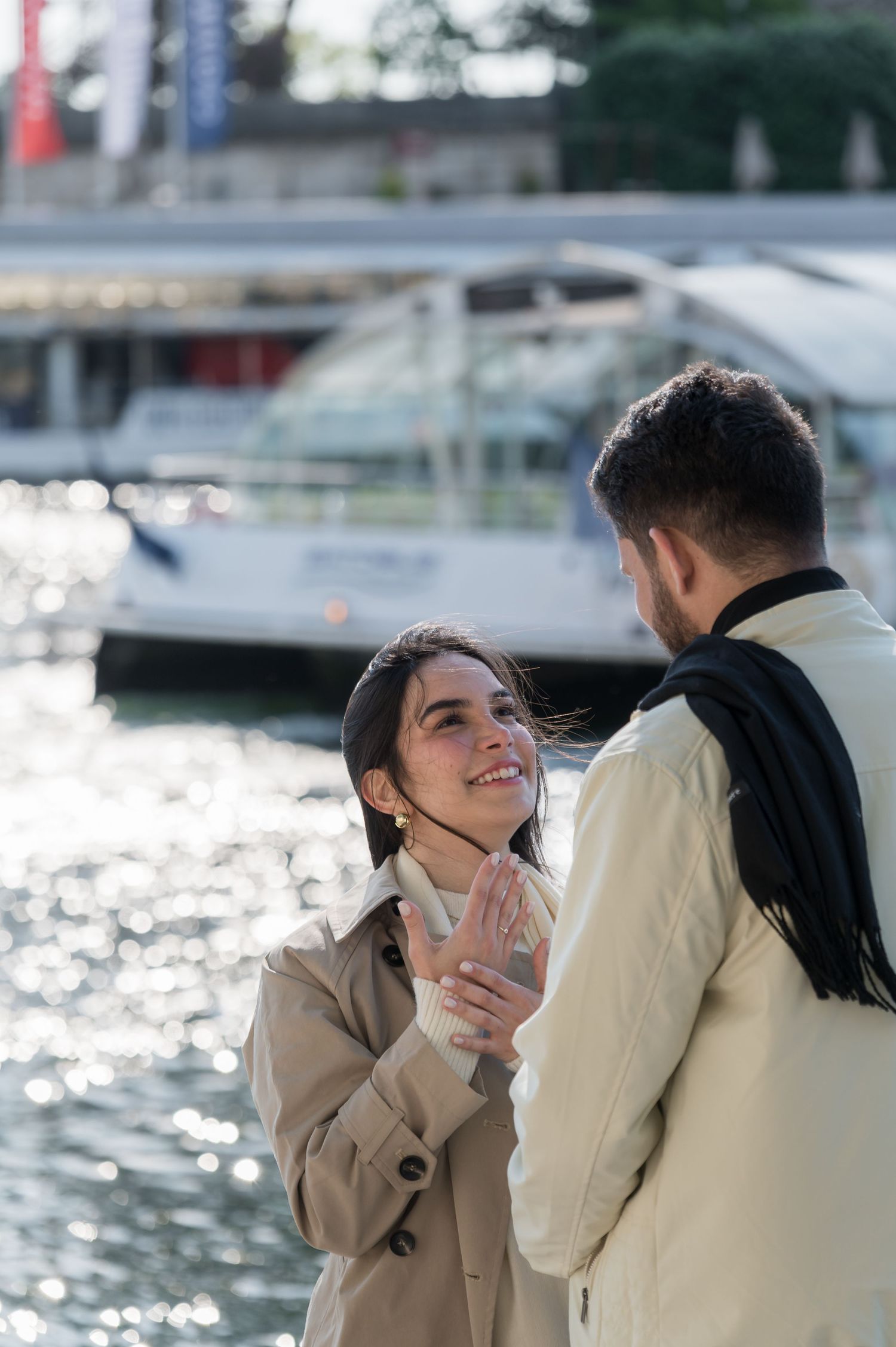 Smiling couple holding hands near a waterfront with a yacht in the background on a sunny day.