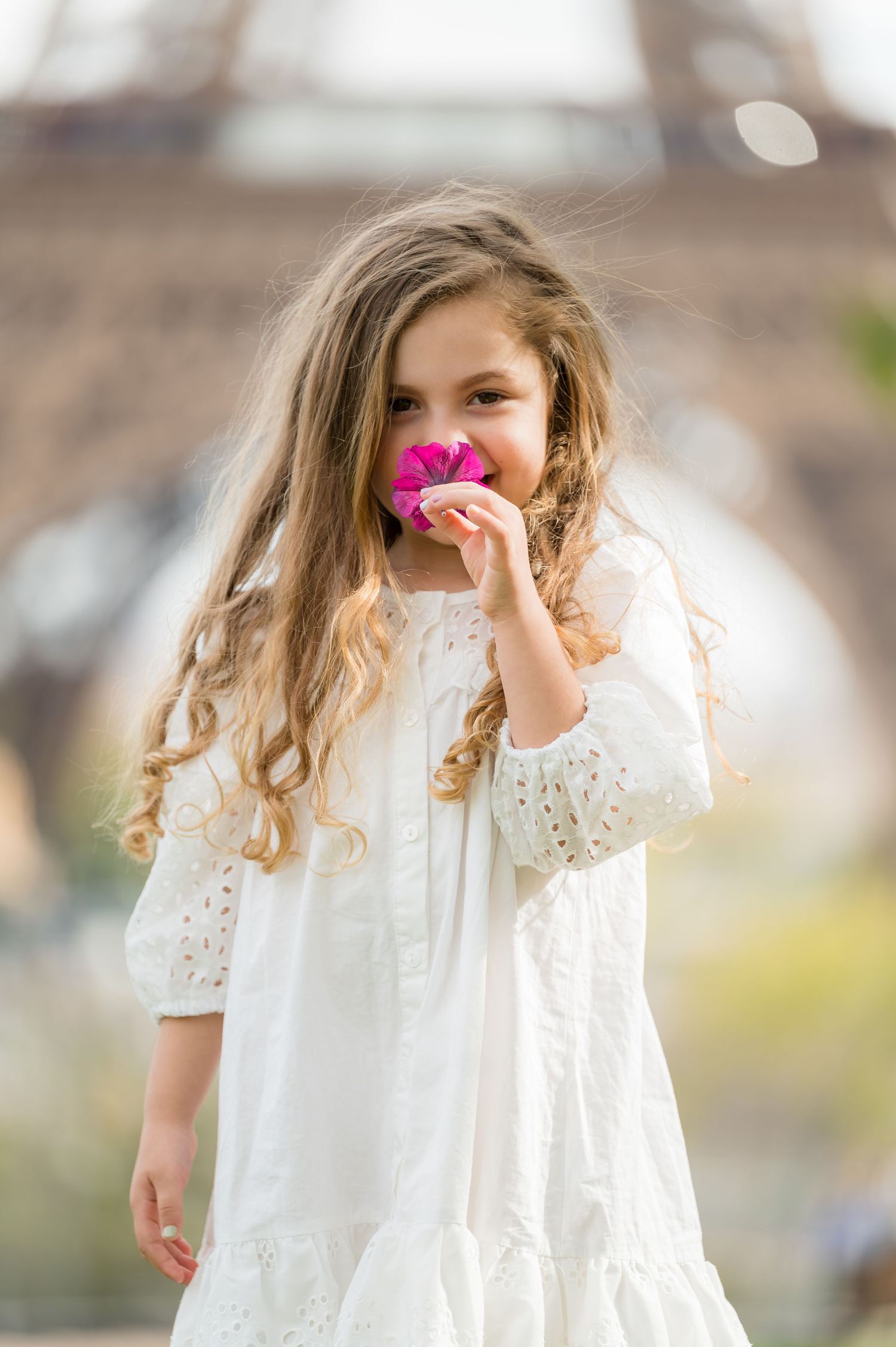 A young child in a white lace dress holds pink flowers while playing in a garden setting.
