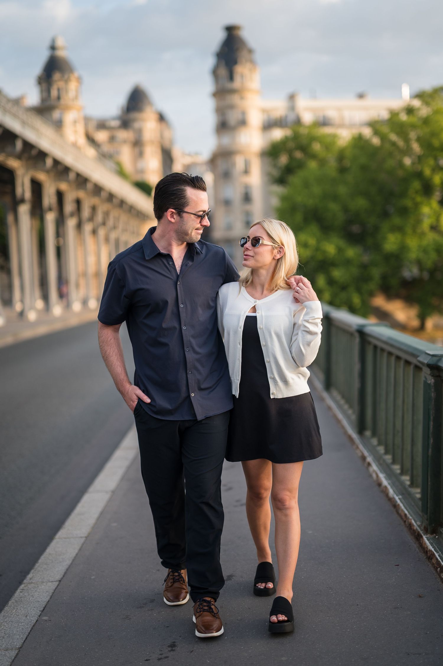 A couple walks together on a bridge with historic architecture visible in the background at sunset.