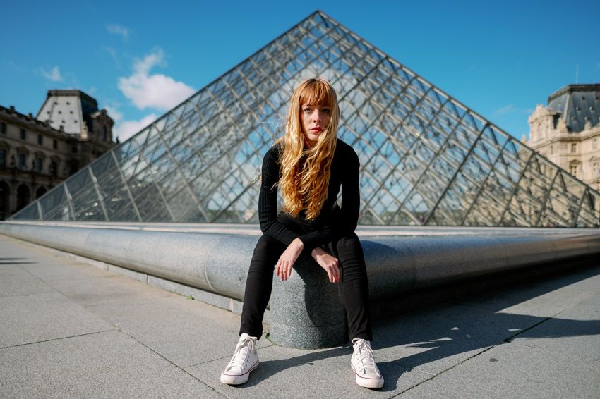 Person in black outfit and white sneakers sits near the iconic glass pyramid at the Louvre Museum in Paris on a sunny day.