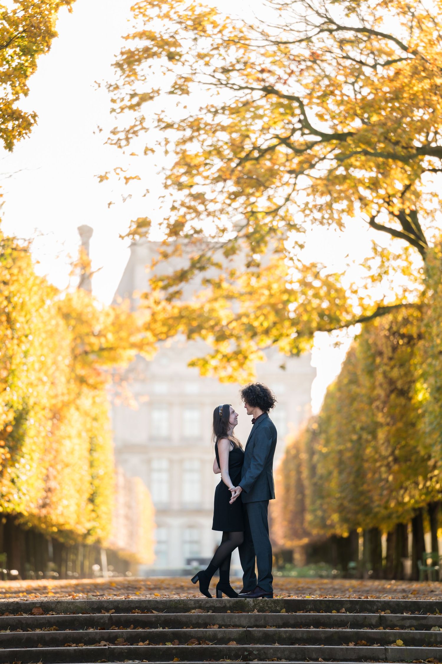 Couple sharing a romantic moment on steps surrounded by golden autumn trees in a Parisian park.
