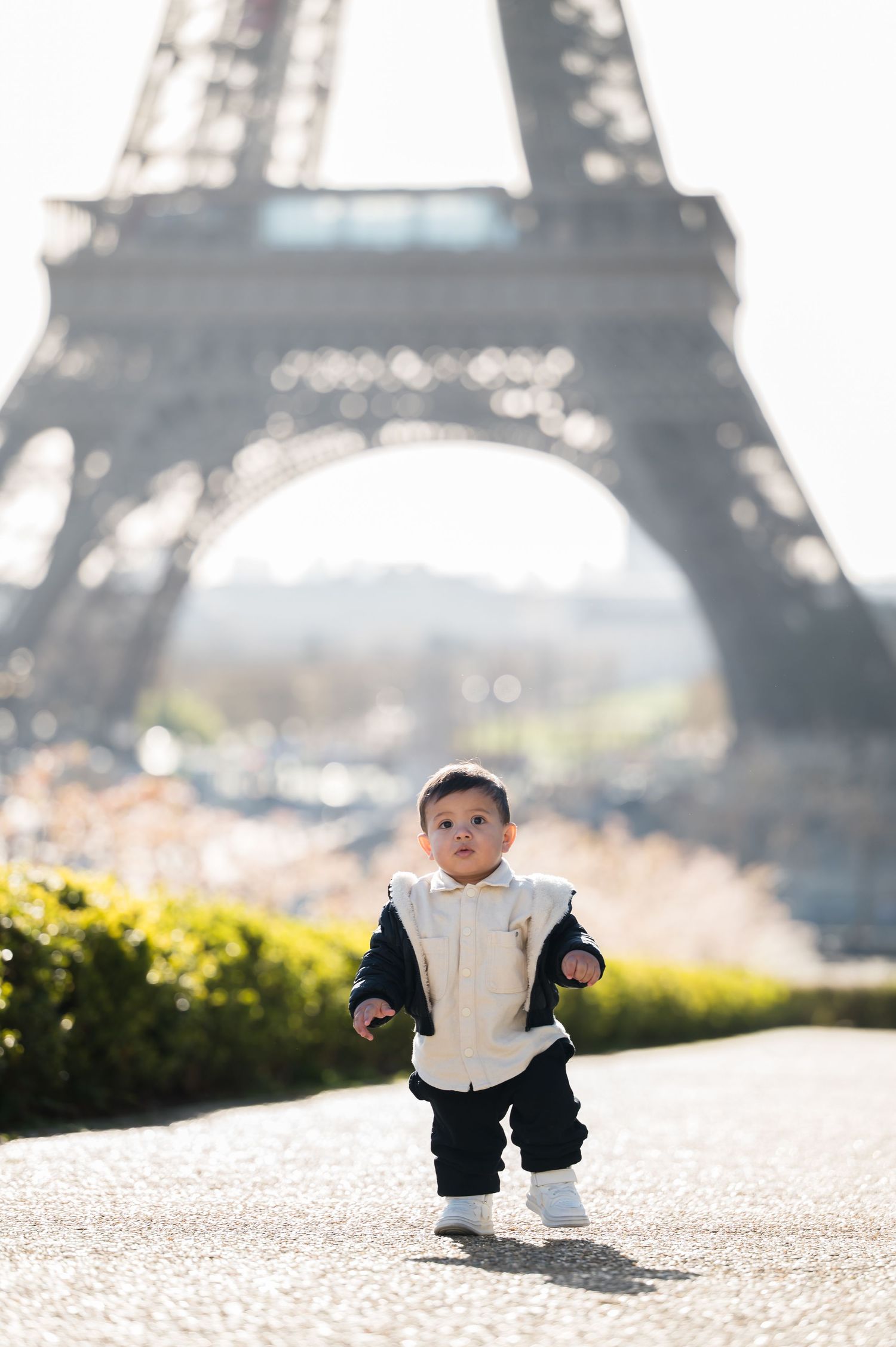 A toddler in a navy outfit walks on a path in front of the iconic Eiffel Tower in Paris, France.