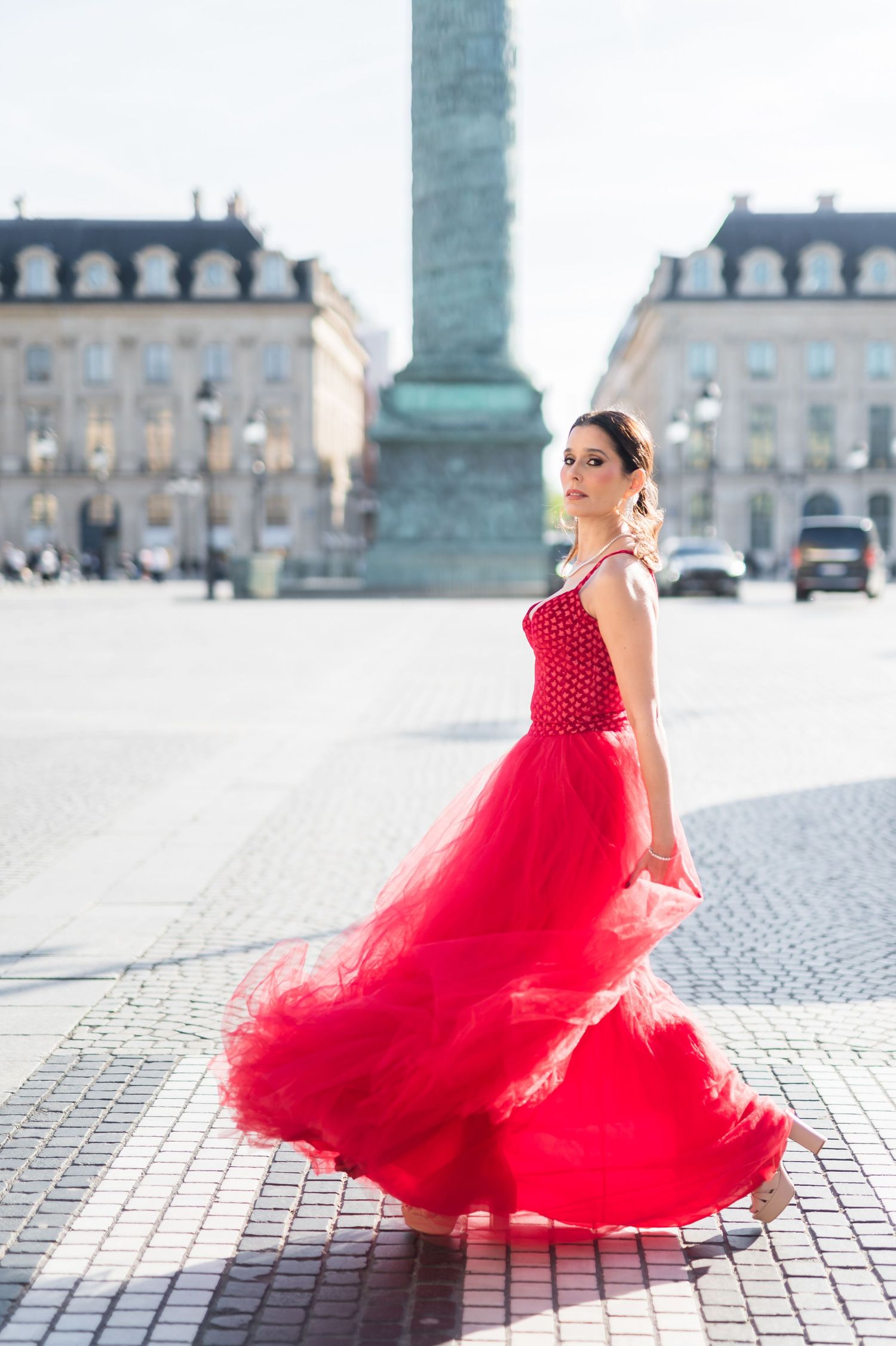 Woman in flowing red tulle gown twirling at Place Vendôme Paris with iconic column in background.