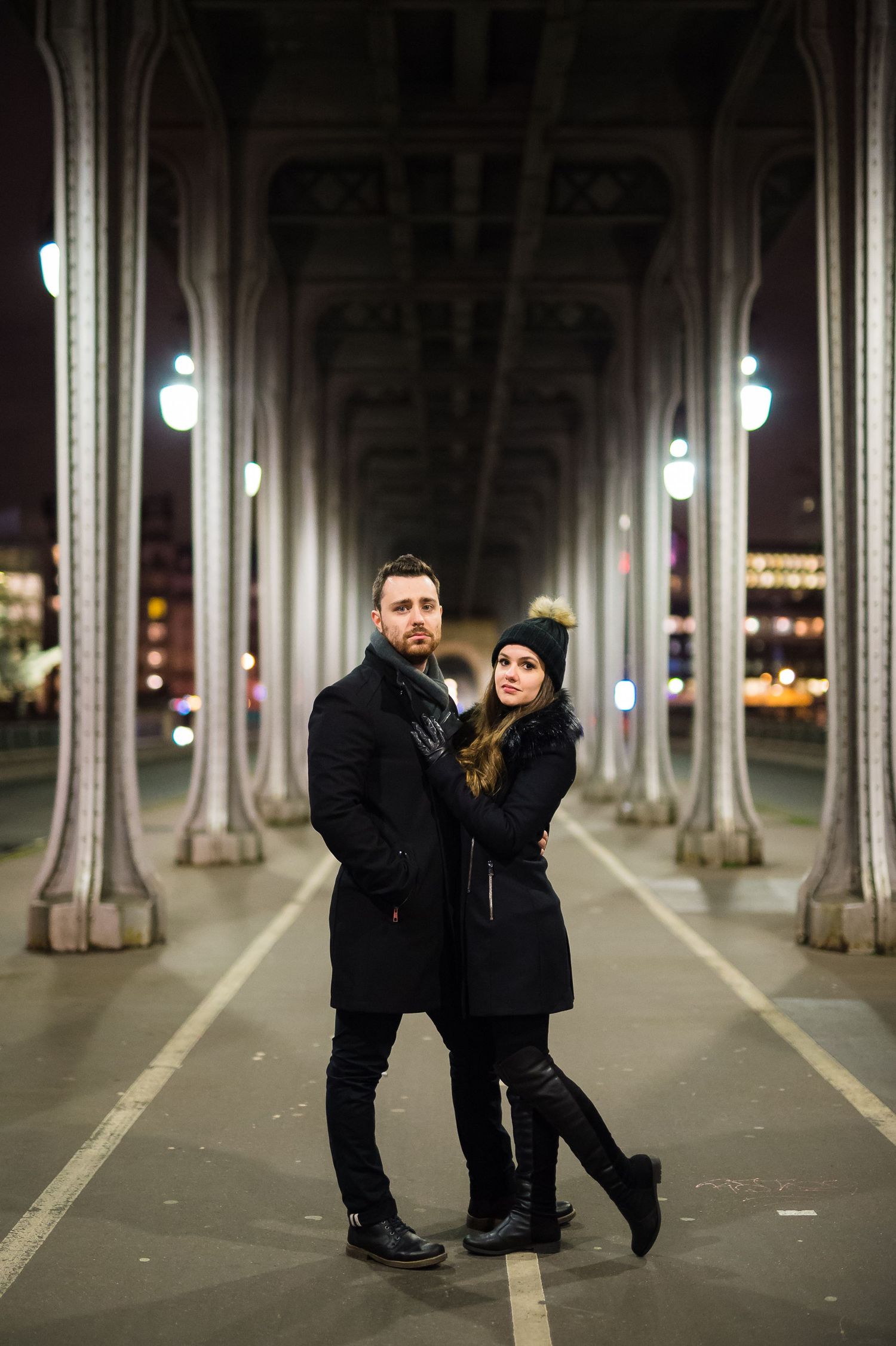 A couple stands beneath the iconic Bir-Hakeim bridge in Paris at night with illuminated columns creating a dramatic perspective.