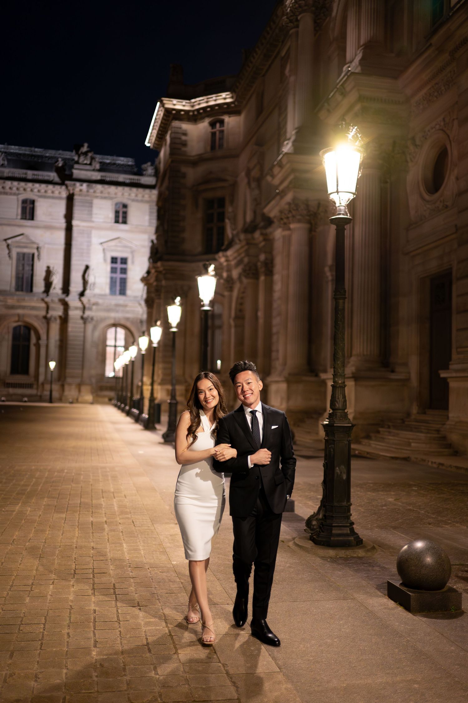 A couple walks arm-in-arm at night along an elegant stone building with ornate lamp posts illuminating their path.