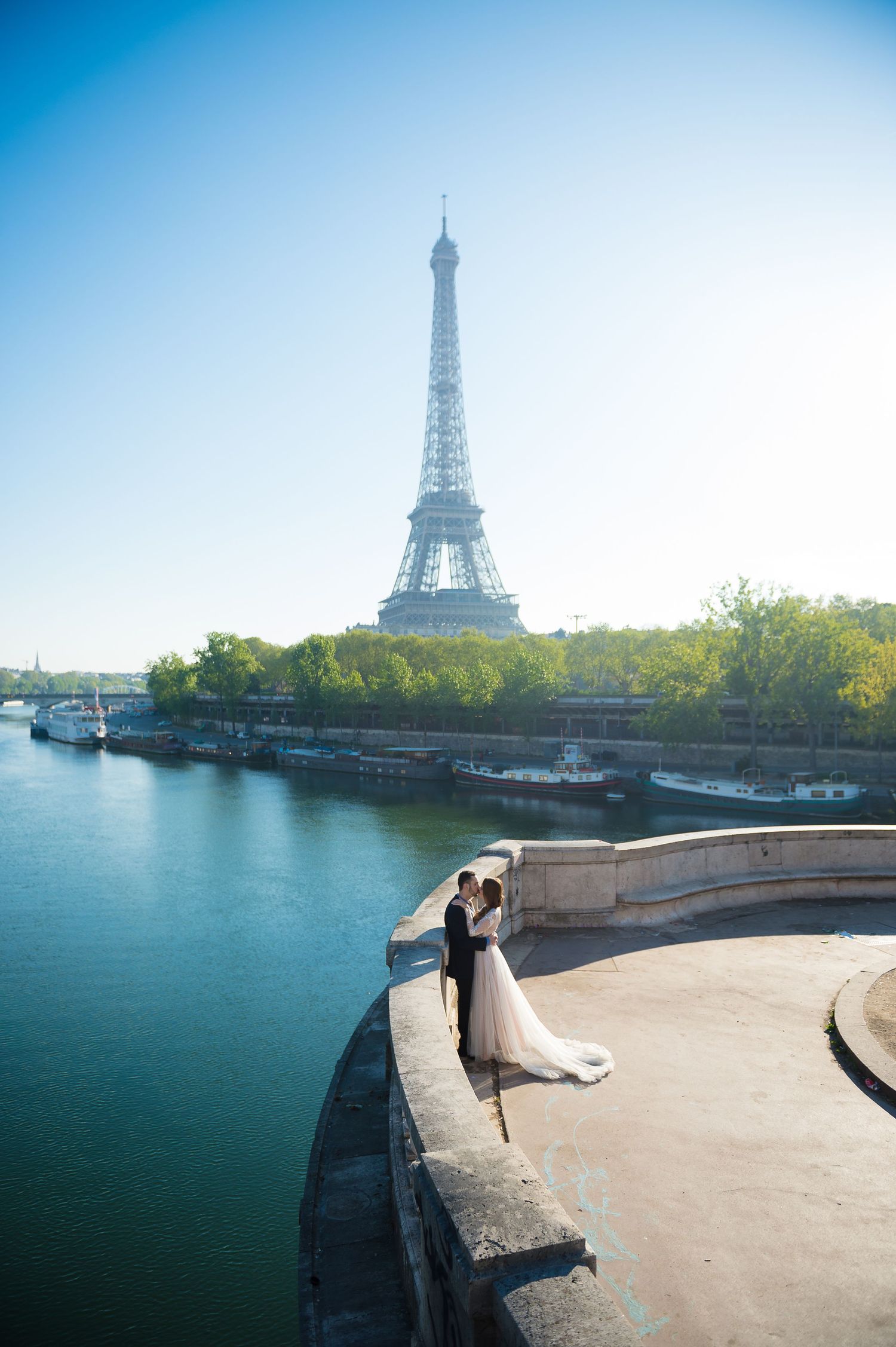 Romantic couple embraced on bridge overlooking Seine River with Eiffel Tower in background on sunny Paris day.