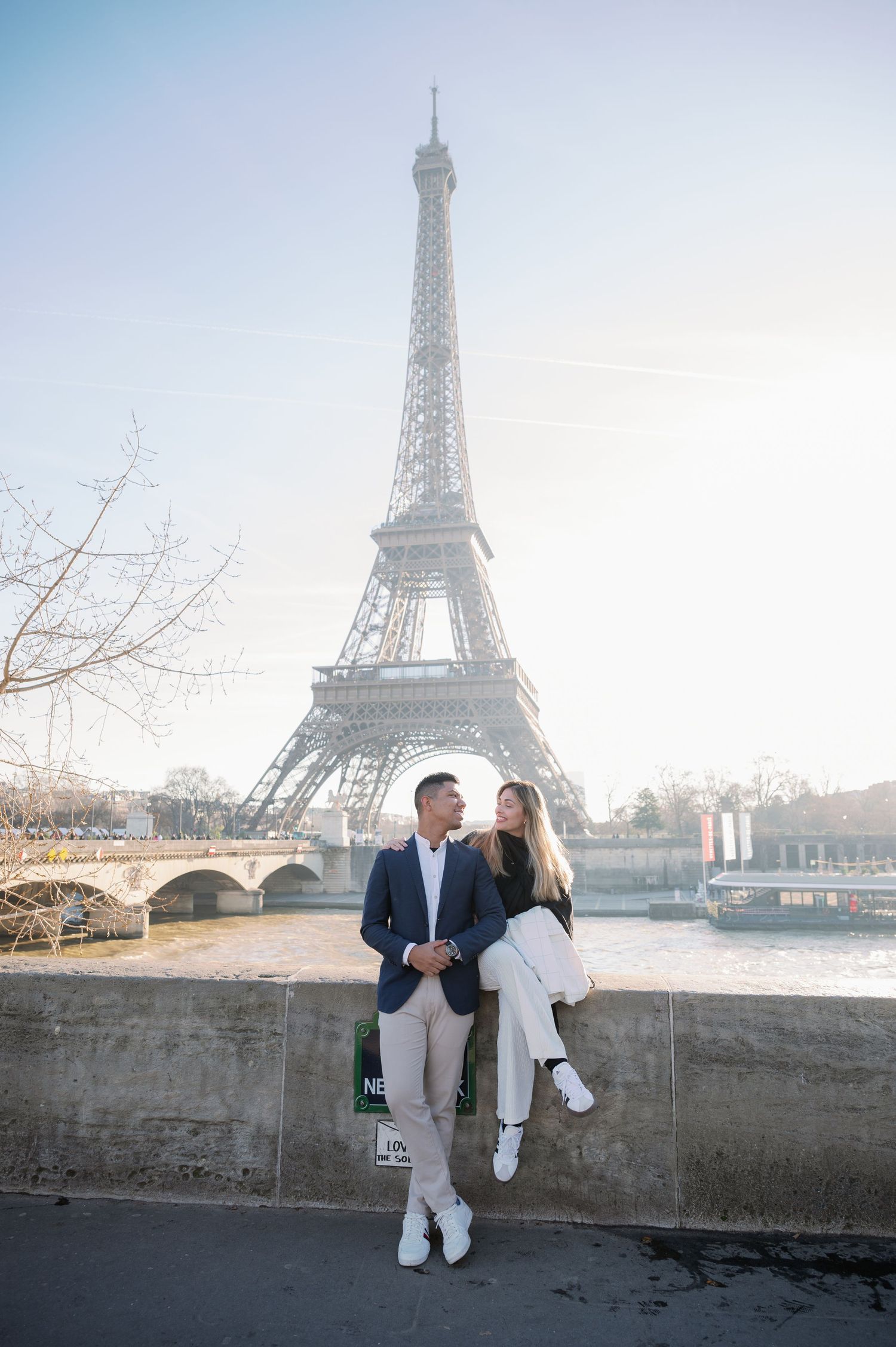 A romantic couple sits on a wall overlooking the Seine with the Eiffel Tower in the background.