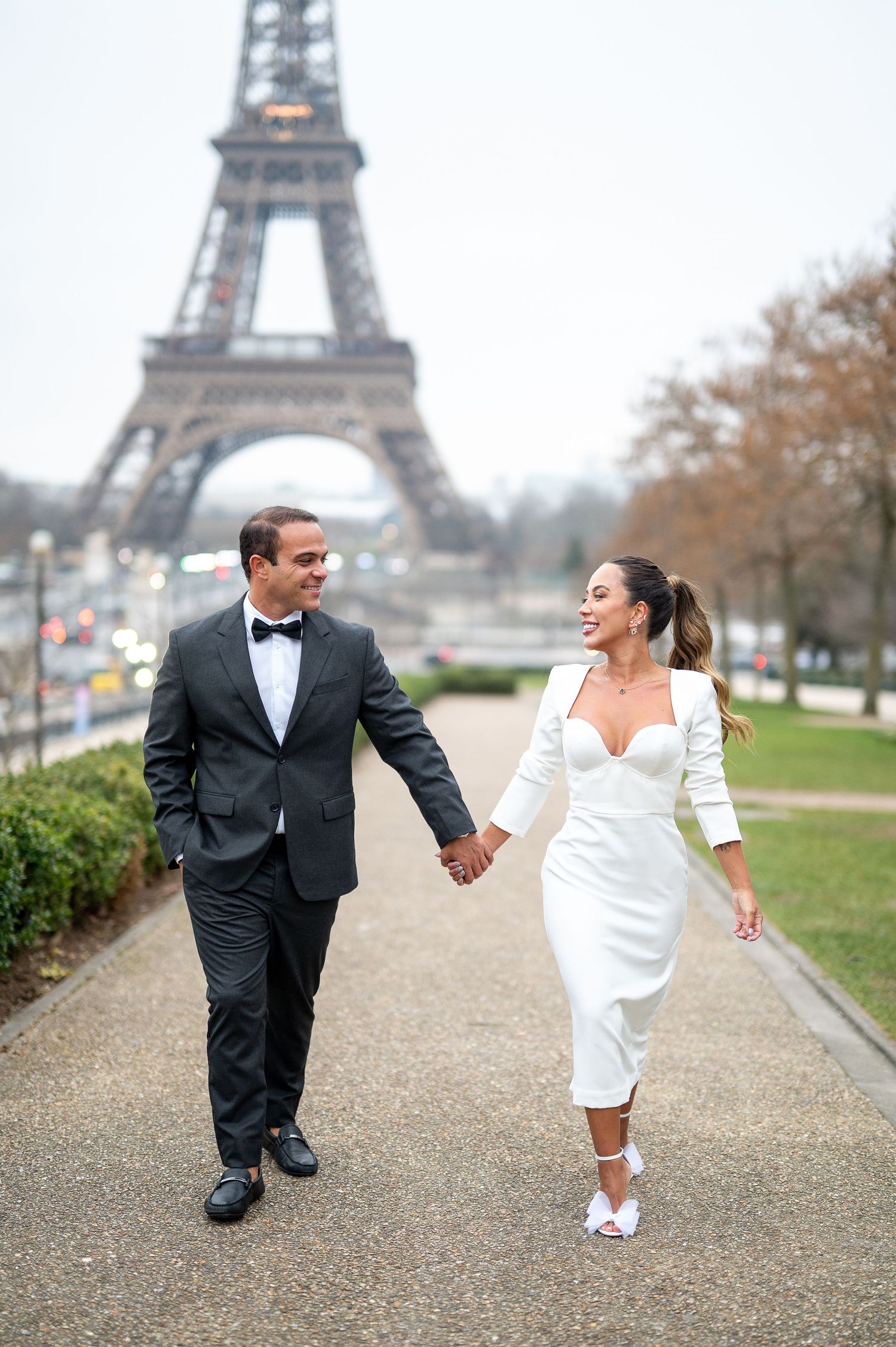 A couple in formal attire walking hand in hand along a path with the Eiffel Tower in Paris as their backdrop.