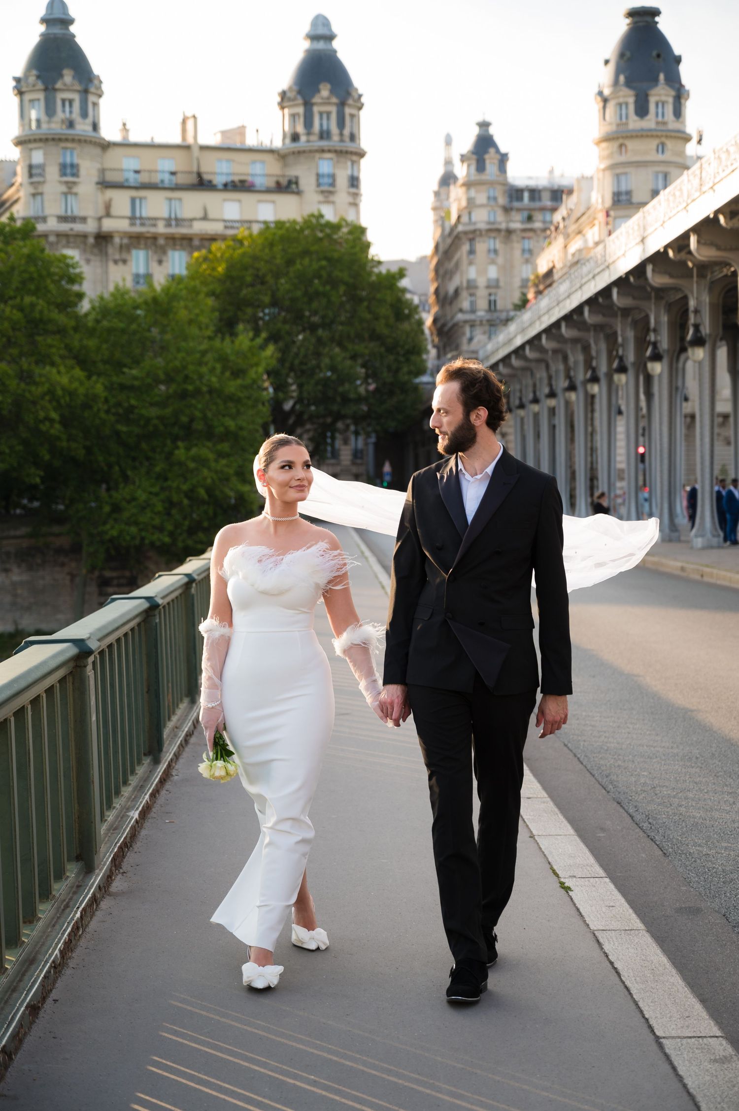 A couple in wedding attire walks along a historic bridge in Paris with elegant architectural buildings in the background.