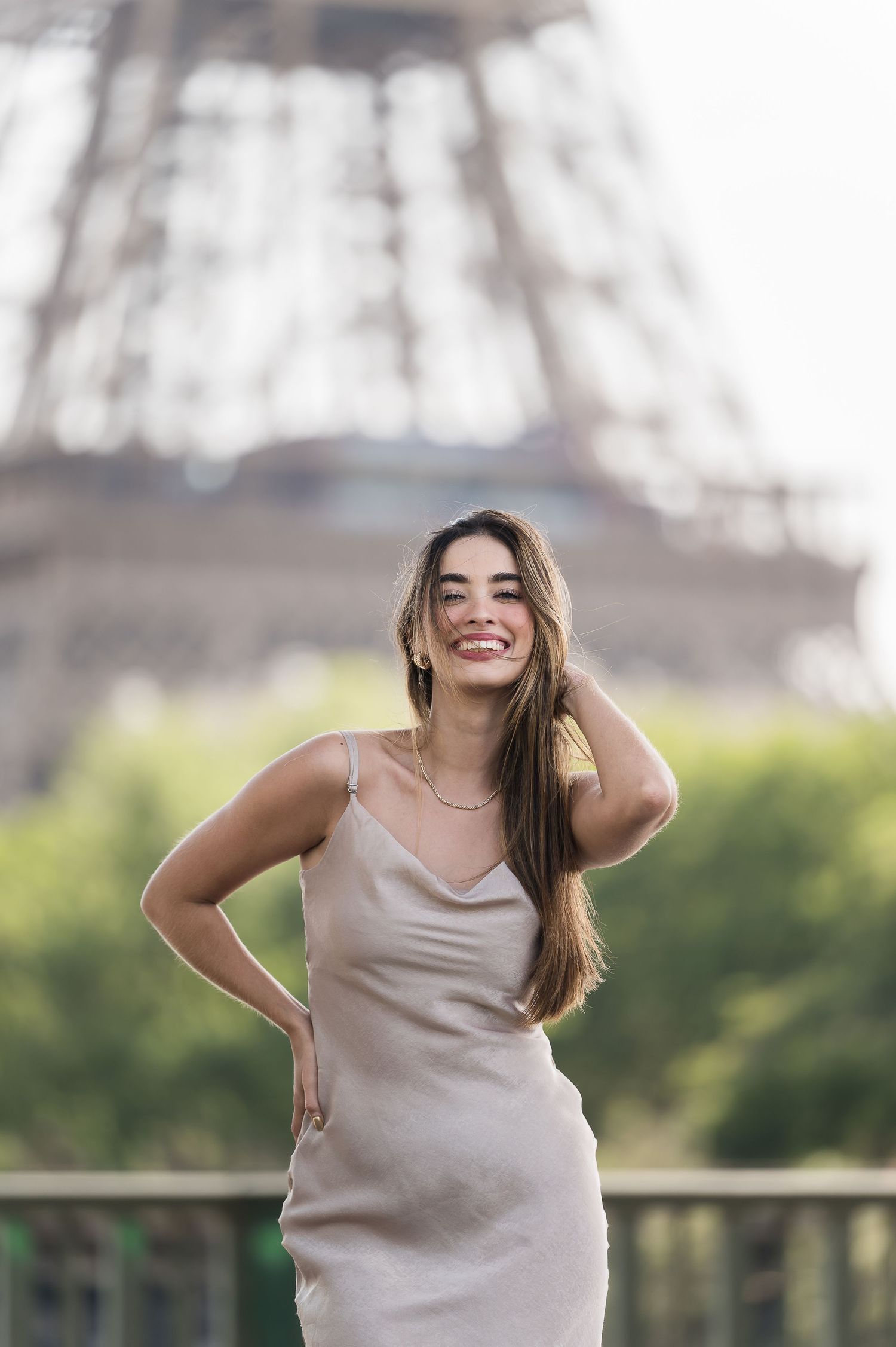Smiling woman in beige slip dress posing in front of the Eiffel Tower in Paris, France.