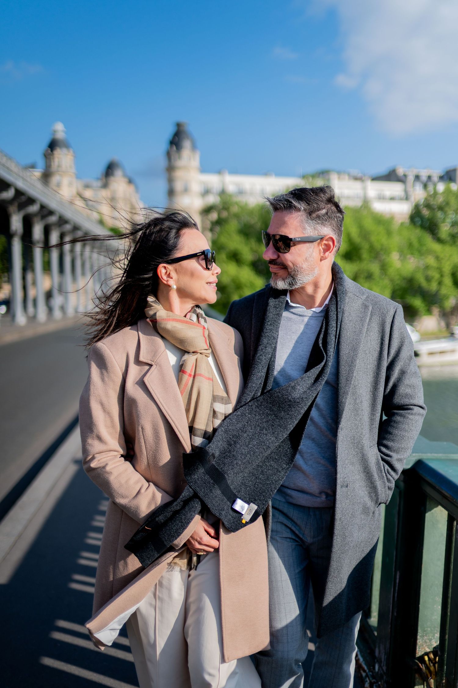 A couple dressed in casual business attire shares a joyful conversation while leaning on a bridge railing in sunny weather.
