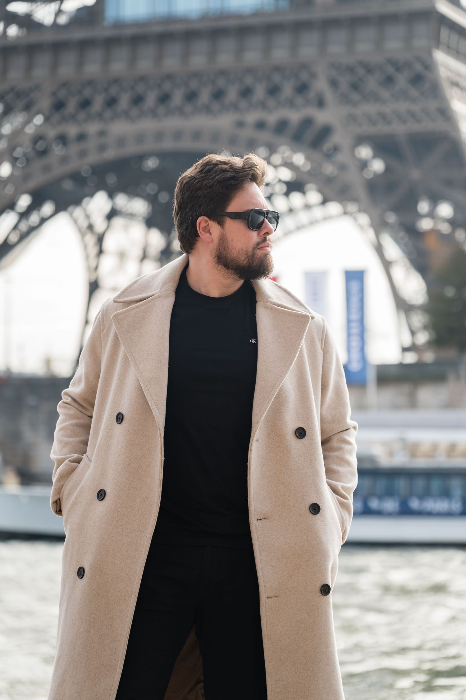 Stylish bearded man in beige coat and sunglasses posing near the Eiffel Tower in Paris.
