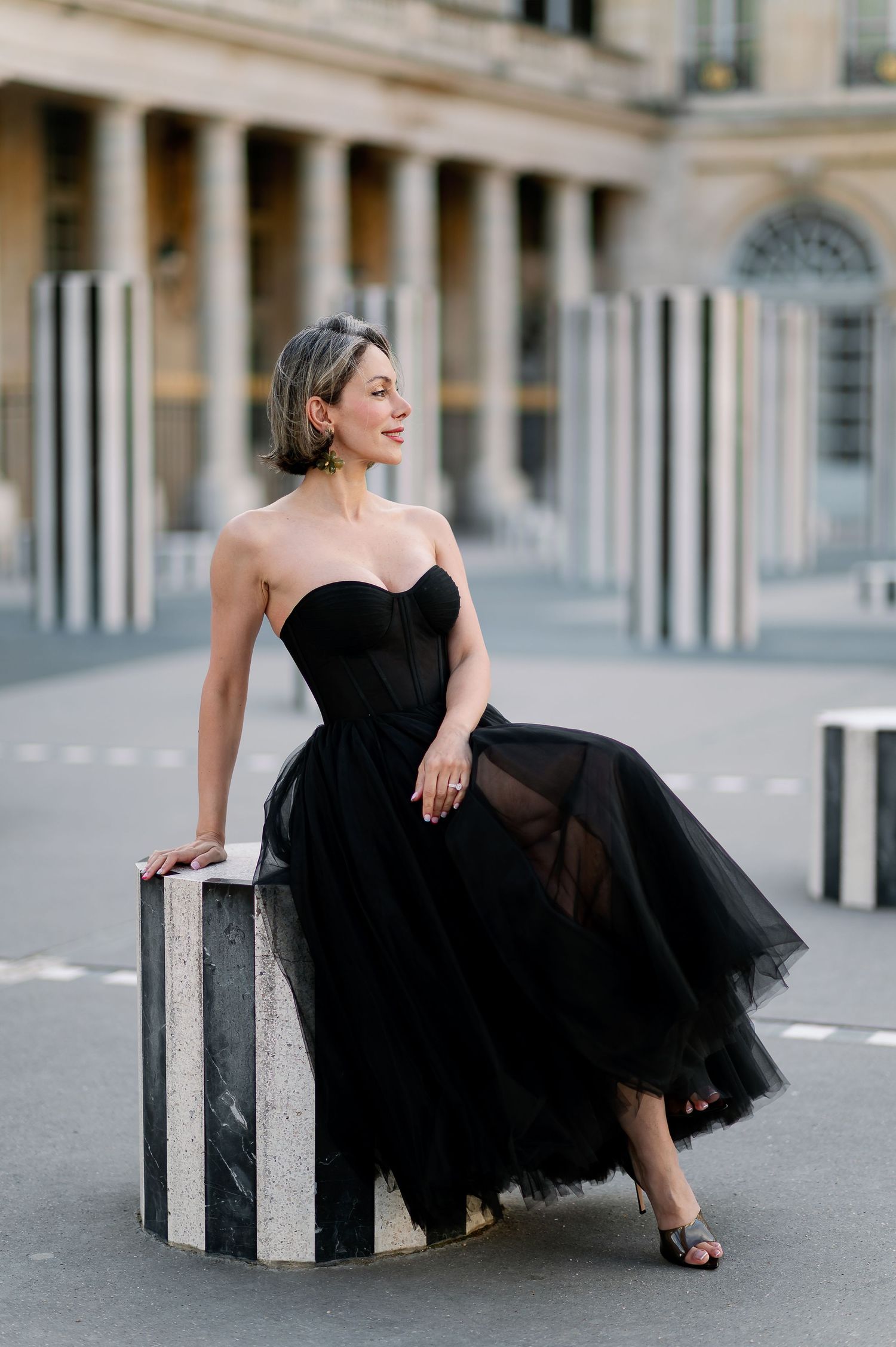 Woman in black strapless tulle gown poses elegantly at Palais Royal, Paris, leaning on striped marble column.