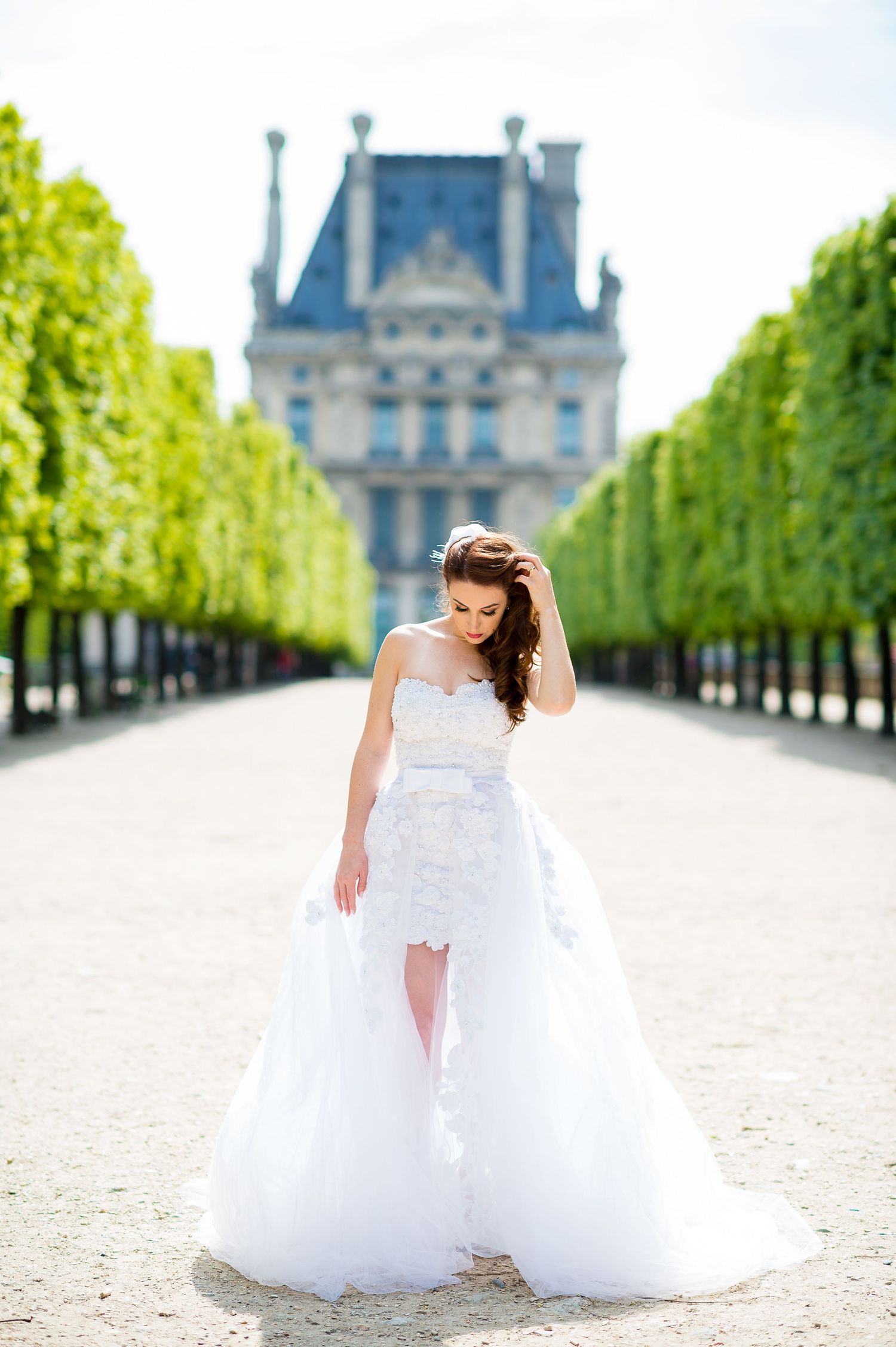 Elegant bride in white lace gown poses along tree-lined path at Tuileries Garden with Louvre Palace in background.