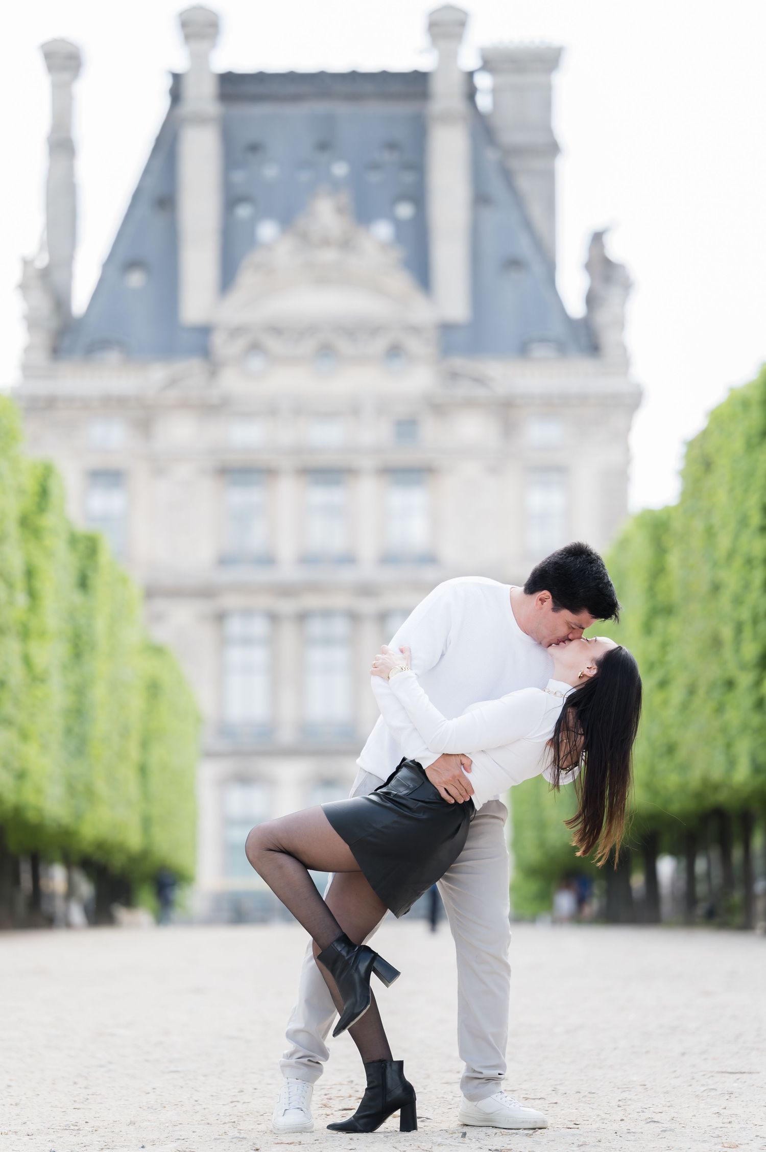 Romantic couple sharing a dip kiss in Tuileries Garden, Paris, with a grand French building in the background.