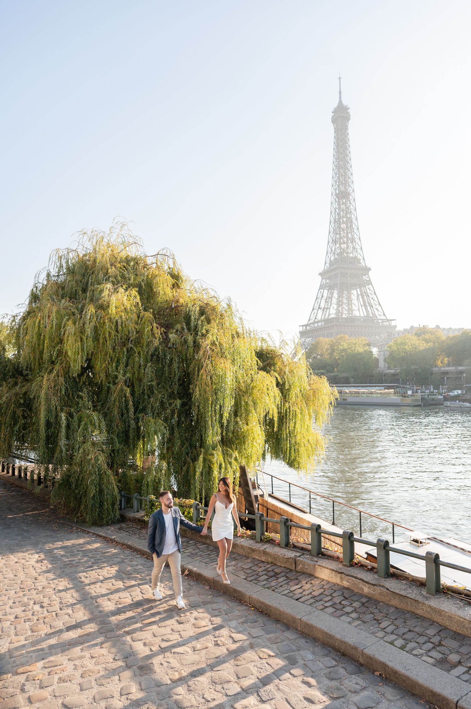 Couple holding hands walking along Seine River with Eiffel Tower and weeping willow tree in sunny Paris background.
