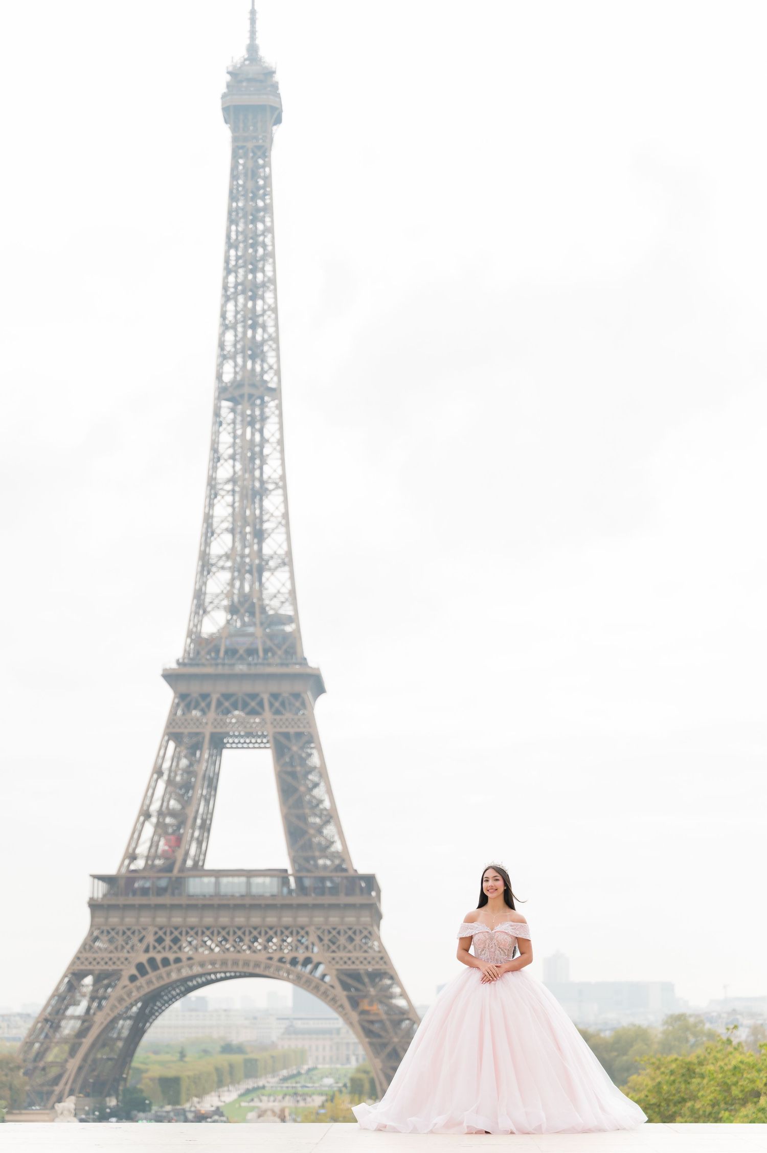A person in a pink ball gown poses in front of the iconic Eiffel Tower in Paris on a cloudy day.