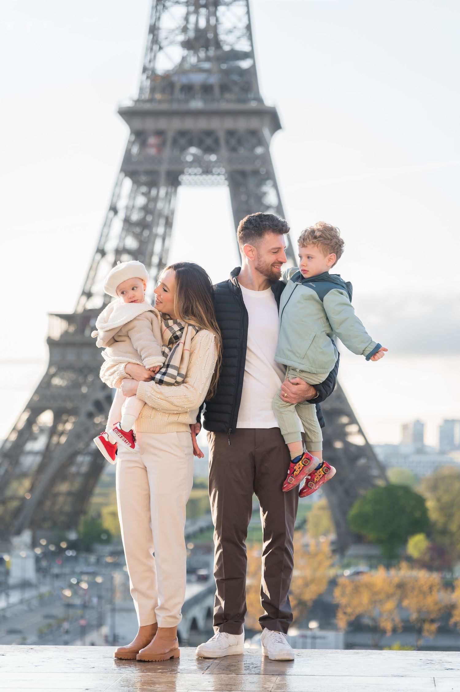 Family of four posing in front of the Eiffel Tower in Paris, parents holding two young children on a sunny day.