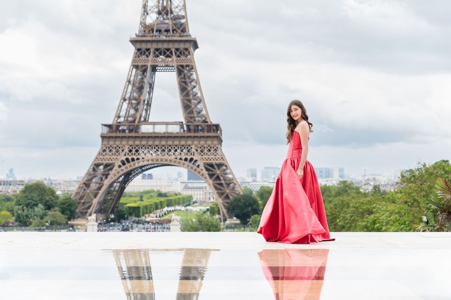 Woman in flowing red dress poses in front of the iconic Eiffel Tower in Paris on a cloudy day.