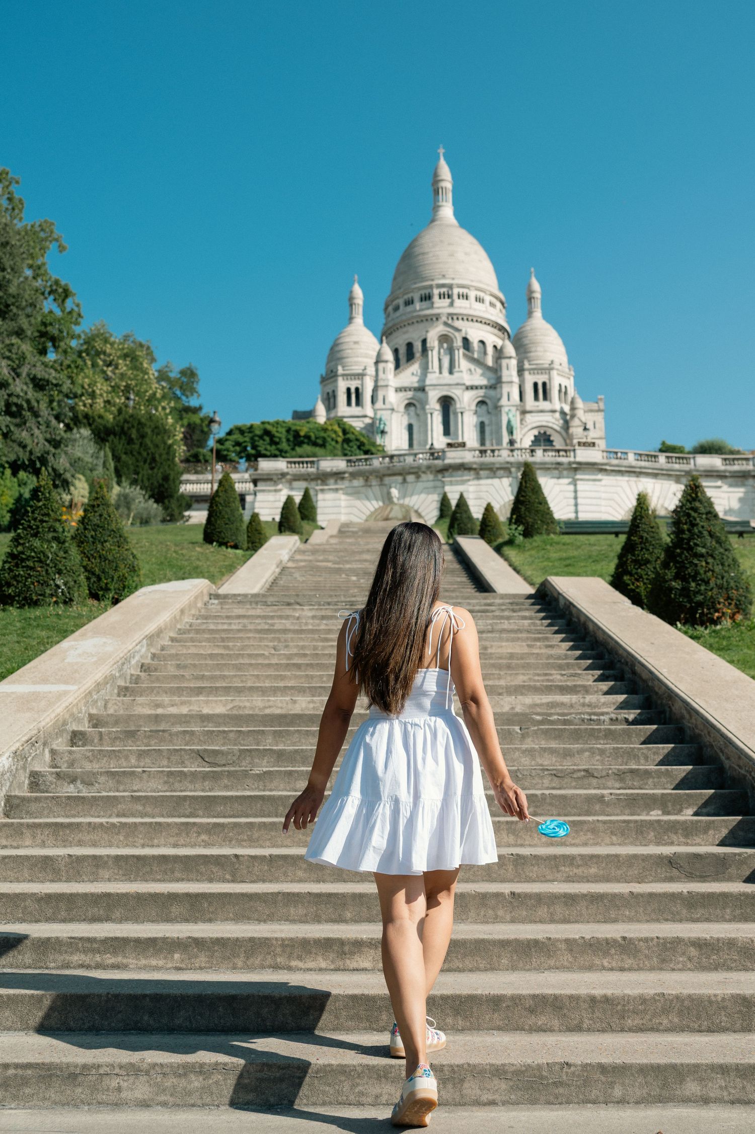 Someone in a white summer dress climbing the steps to the iconic Sacre-Coeur Basilica in Paris on a sunny day.